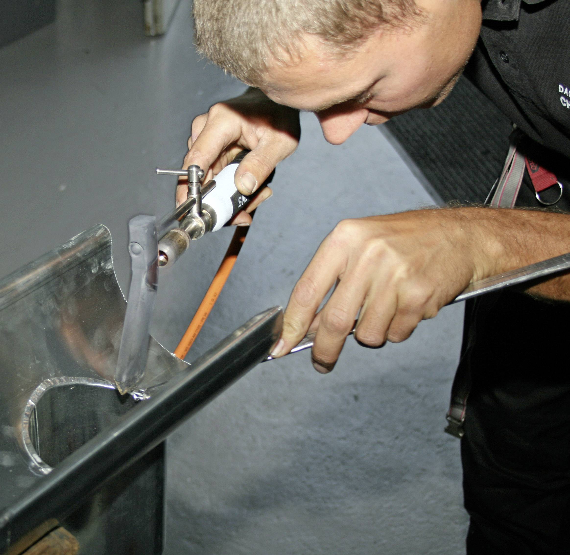 Ein Mann schweißt mit einem Schweißbrenner ein Metallstück auf einem Tisch. Seine Augen sind auf die Arbeit konzentriert.