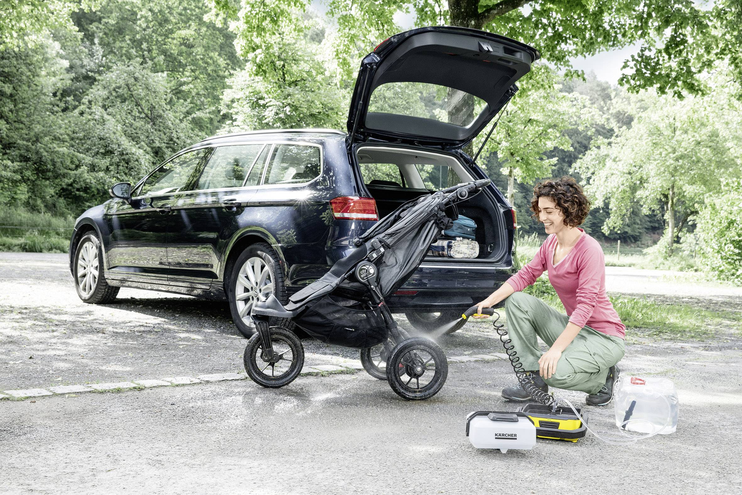 Eine Person spritzt einen Kinderwagen neben einem Auto mit einem Hochdruckreiniger ab. Die Kofferraumklappe des Autos ist geöffnet.