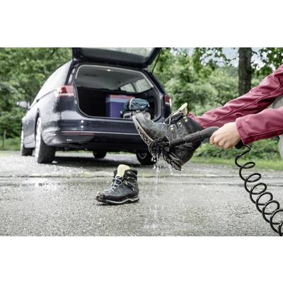 Person spritzt Wanderstiefel mit Wasser ab neben geparktem Auto, Kofferraum offen mit Ausrüstung. Outdoor-Aktivität oder Reisevorbereitung.