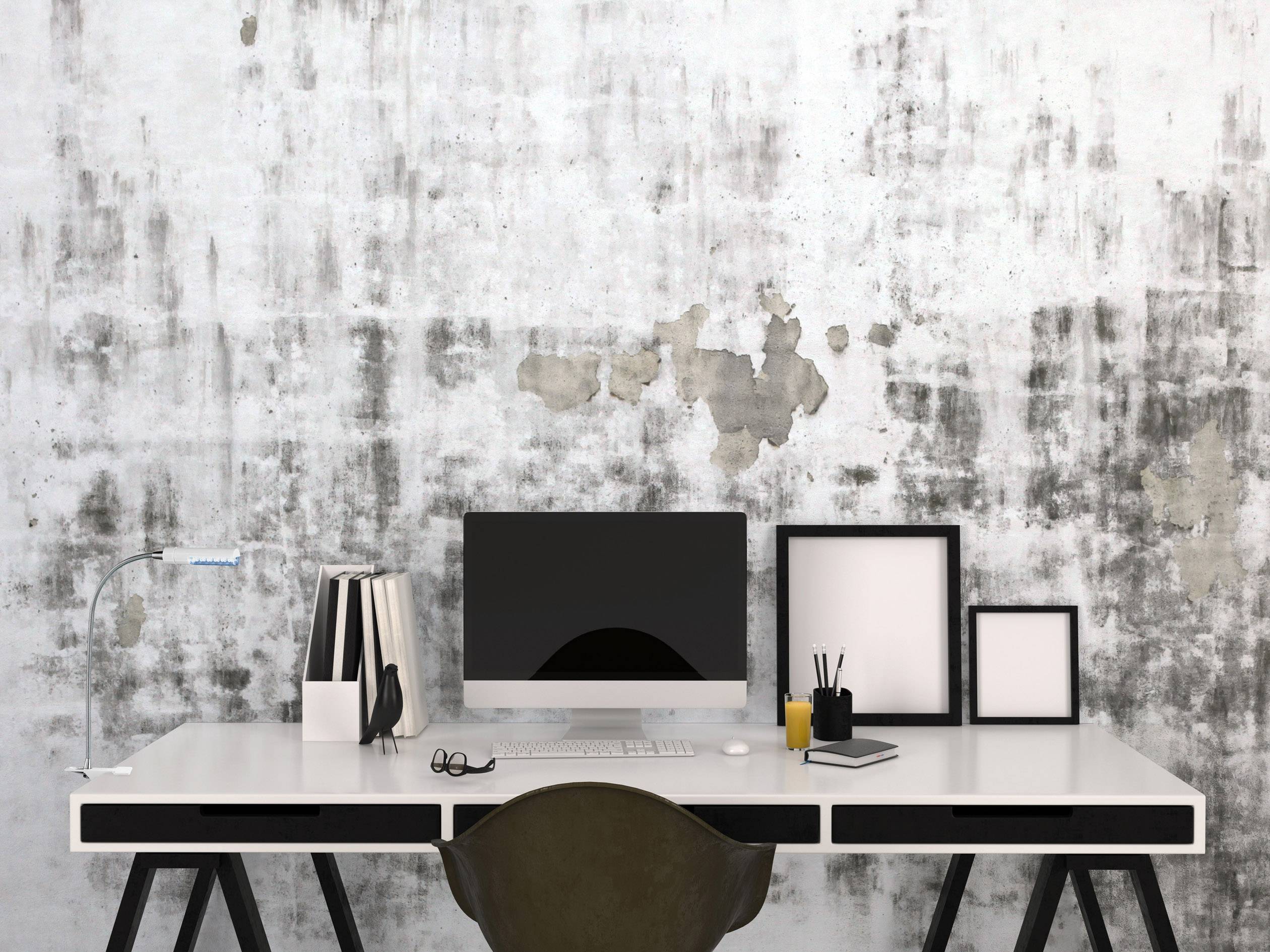 Ein minimalistisch eingerichteter Schreibtisch mit einem Computer, Büchern, Bildern und Schreibwaren vor einer betonartigen Wand.