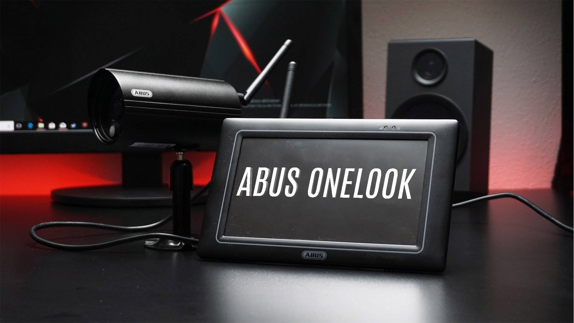 Überwachungskamera und Monitor auf Tisch. Auf dem Monitor steht 'Abus OneLook'. Im Hintergrund ein Computer und Lautsprecher.