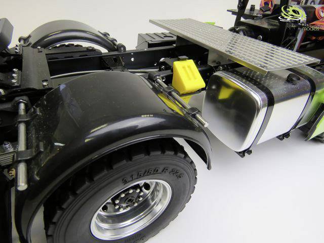Nahaufnahme eines Modell-Lkw-Chassis mit sichtbaren Kotflügeln, einem glänzenden Tank, gelben Komponenten und einer Metallplatte.
