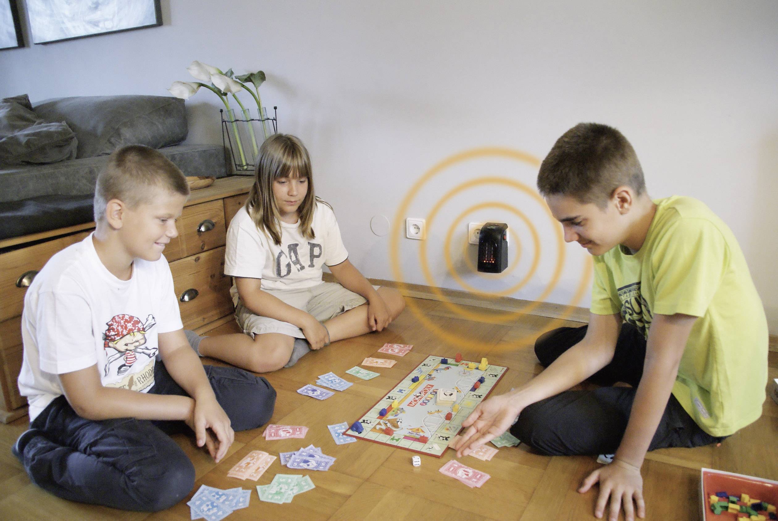 Drei Kinder spielen ein Brettspiel auf dem Boden. Um sie herum liegen bunte Geldscheine. An der Wand ist ein schwarzer Router mit Signalwellen.
