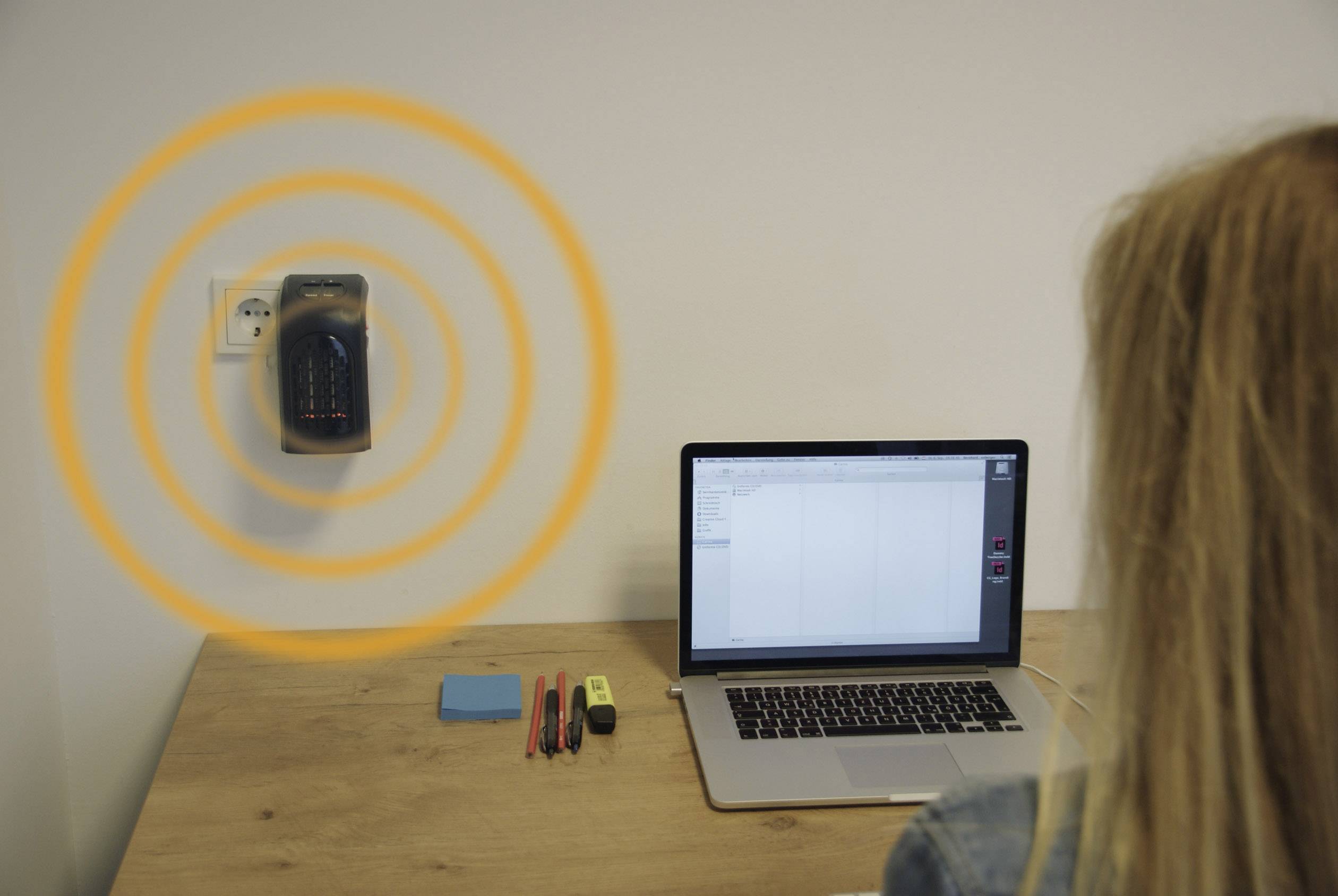 Eine Person sitzt an einem Tisch mit einem Laptop. An der Wand ist ein Gerät mit konzentrischen orangen Kreisen, die Funkwellen darstellen.