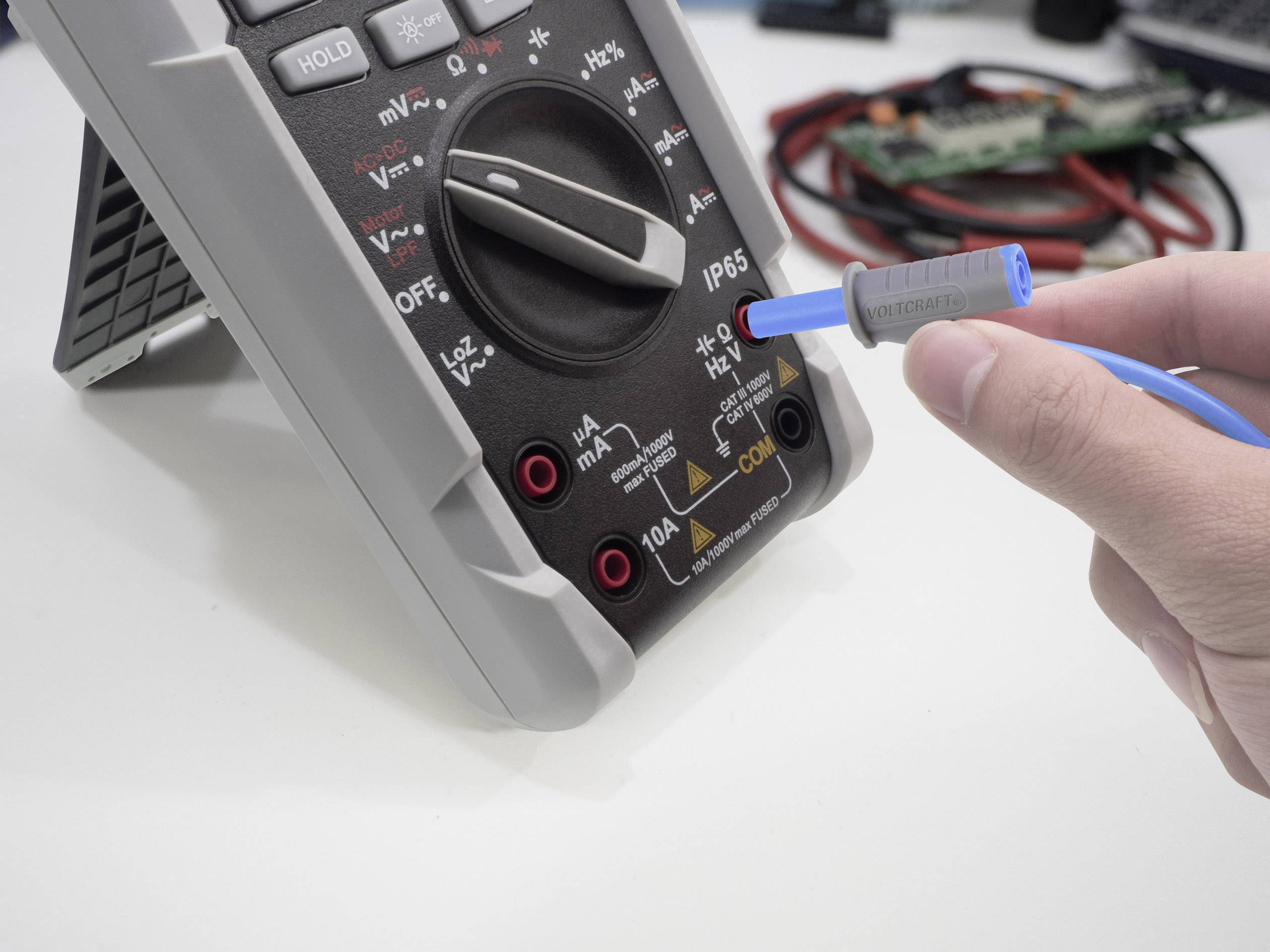 Eine Person verwendet ein digitales Multimeter auf einem Tisch. Die Skala zeigt verschiedene Messbereiche. Ein blaues Kabel wird angeschlossen.