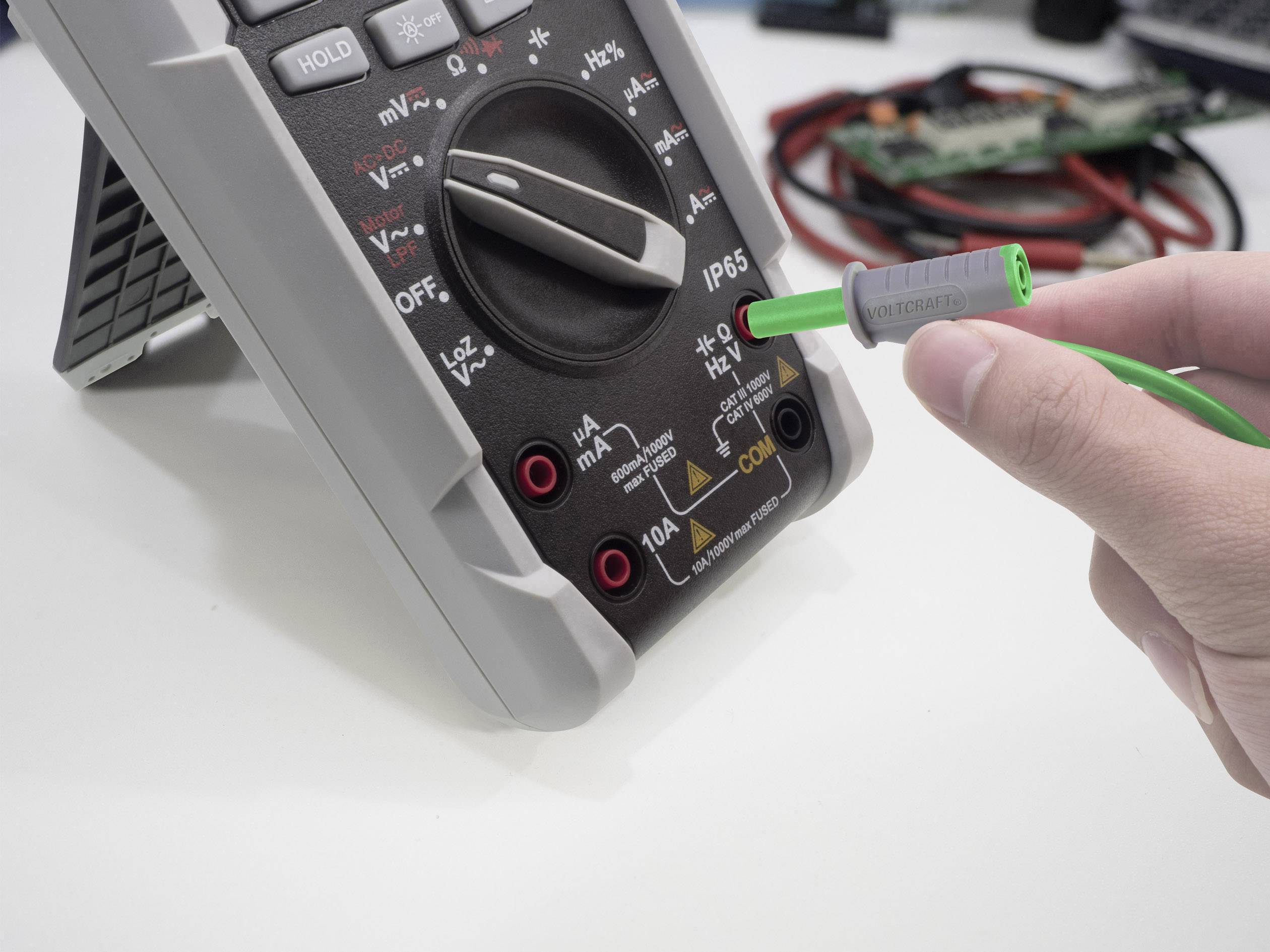 Ein Multimeter wird von einer Hand verwendet. Eine grüne Sonde wird an den Anschluss des Multimeters angeschlossen. Im Hintergrund sind Kabel sichtbar.