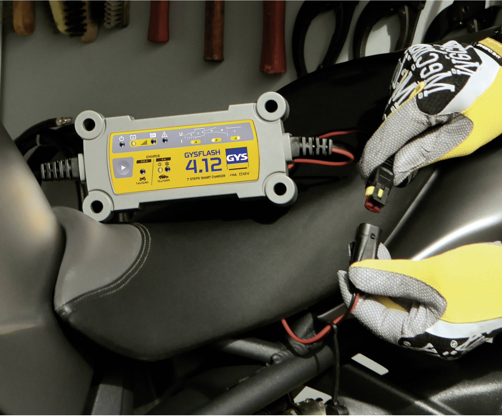 Ein Batterieladegerät liegt auf einem Motorradsitz, umgeben von Schraubenschlüsseln in einer Werkstattumgebung.