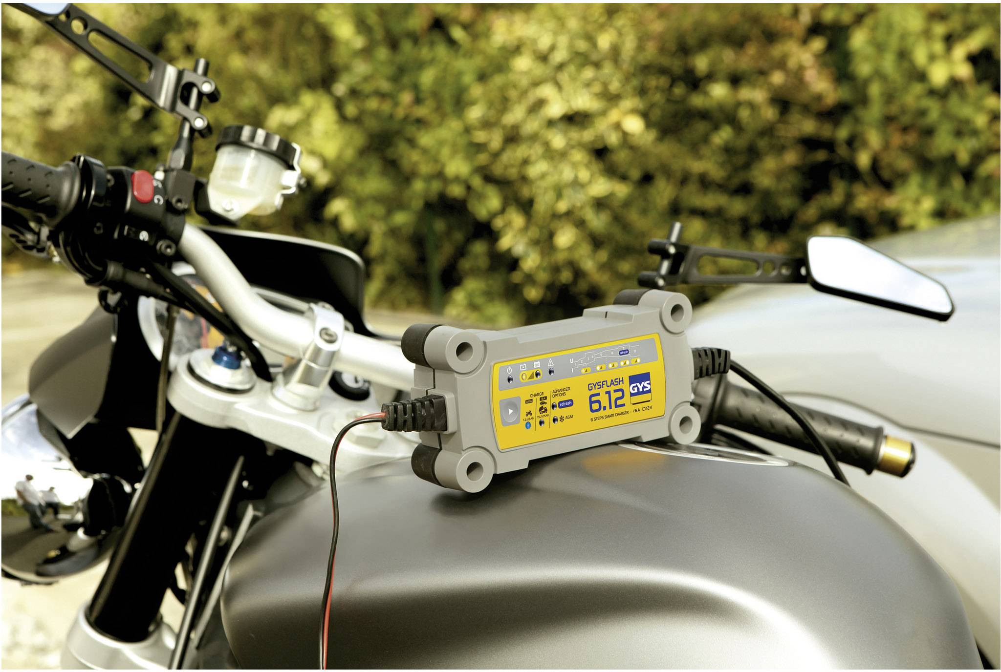 Ein Batterieladegerät ist auf einem Motorrad angebracht, das in einem Freien geparkt ist. Das Gerät zeigt eine Spannung von 6,12 Volt an.