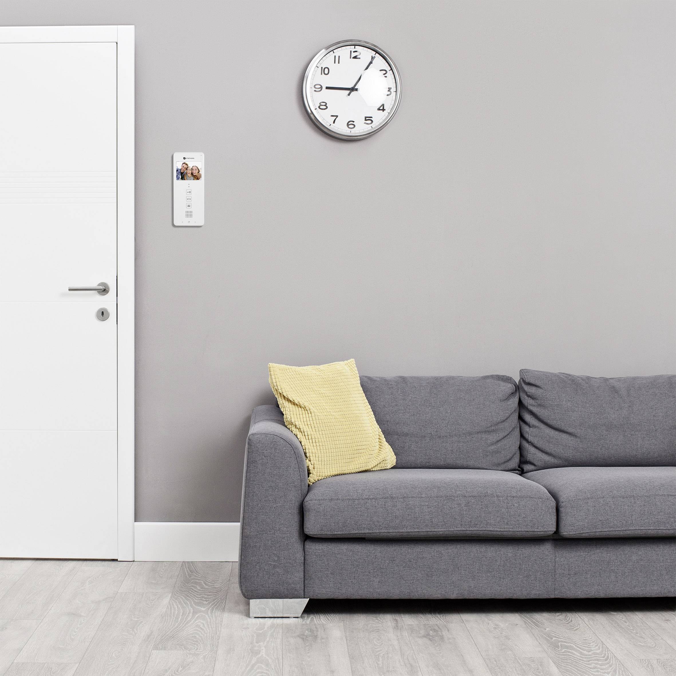 Ein graues Sofa mit zwei Kissen vor einer hellen Wand. Rechts hängt eine Uhr, links ein Thermostat.