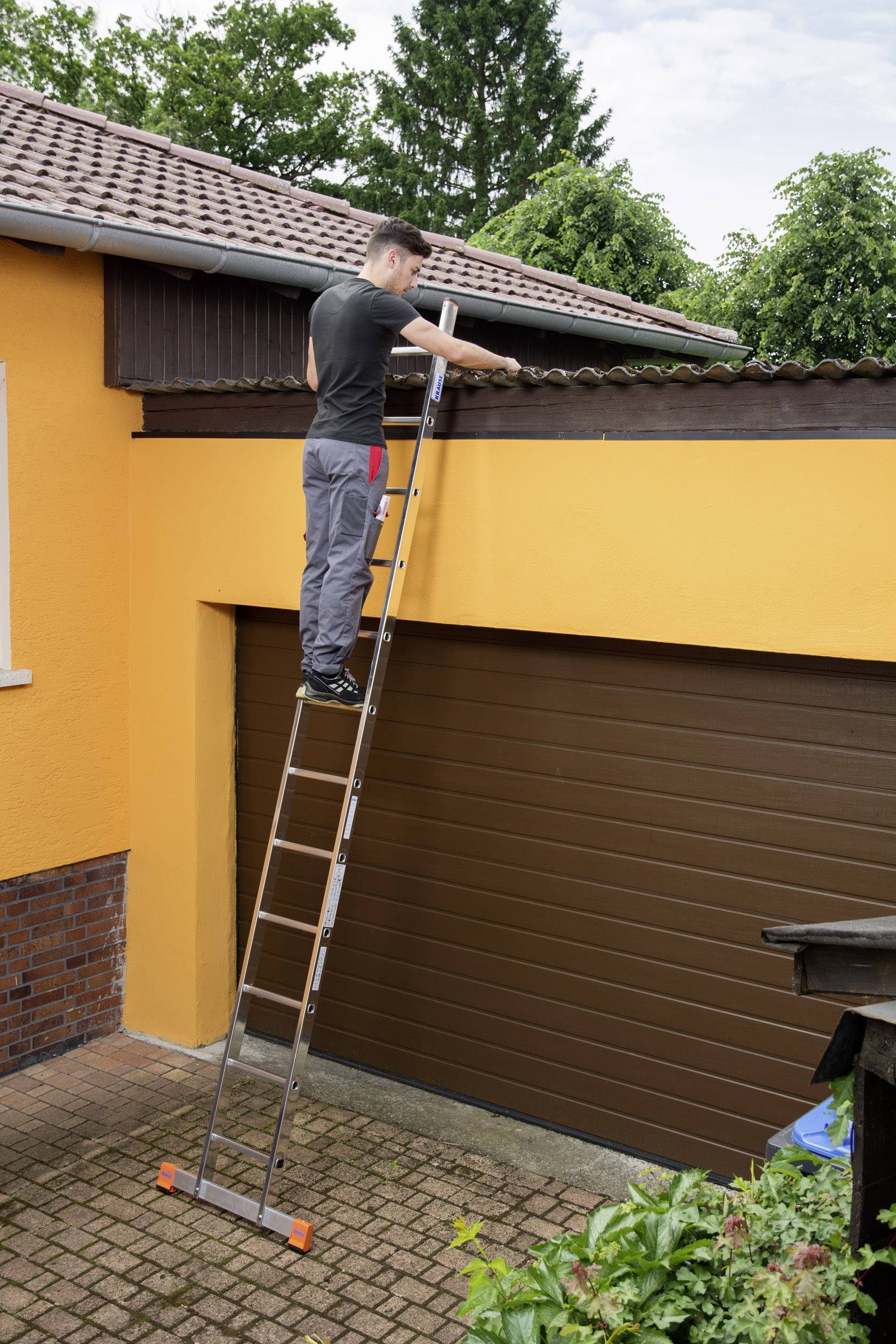 Ein Mann steht auf einer Leiter und repariert das Dach eines gelben Hauses. Im Hintergrund sind Bäume zu sehen.