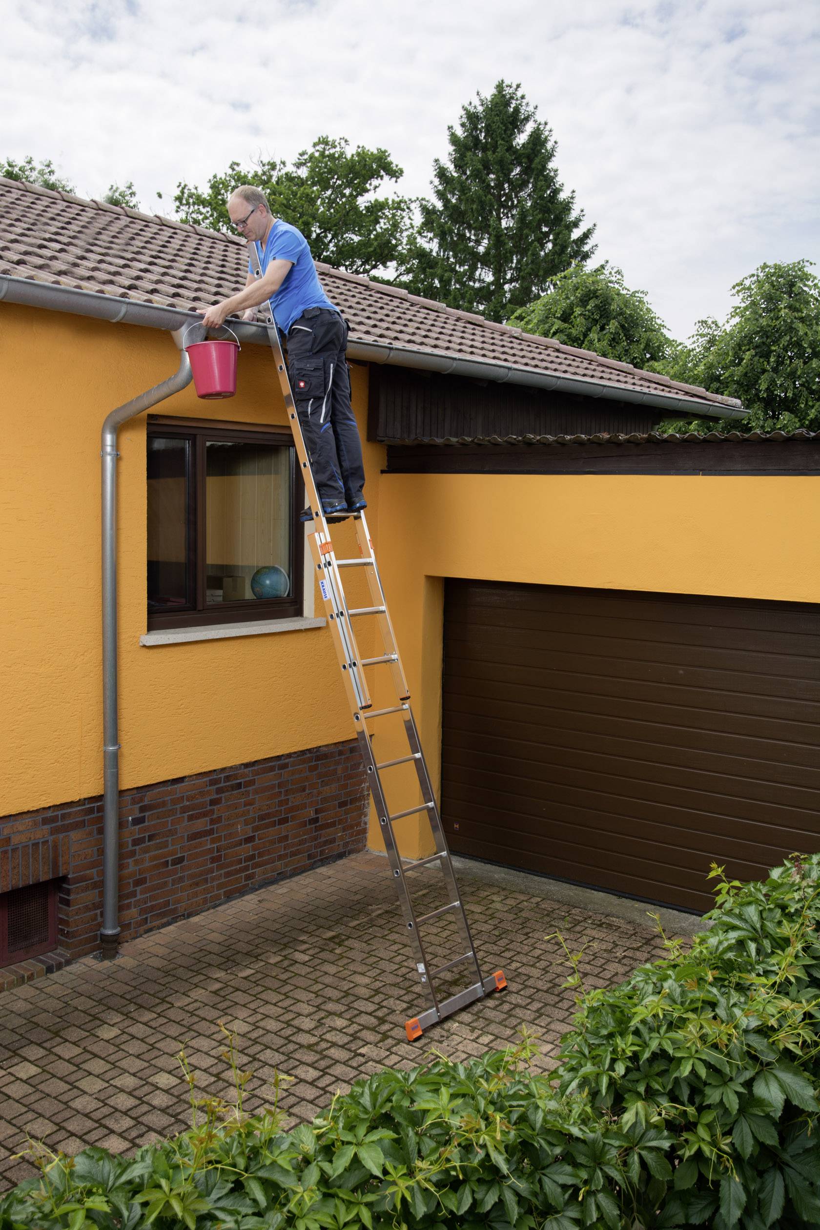 'Eine Person steht oben auf einer Leiter und reinigt die Dachrinne eines Hauses mit einem roten Eimer.'