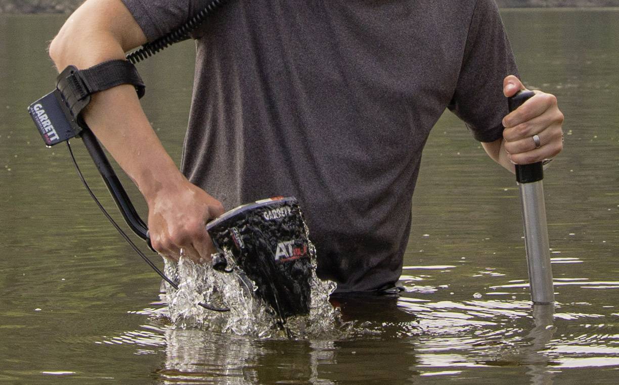Eine Person steht bis zur Hüfte in Wasser und bedient ein Metalldetektorgerät, das im Wasser nach Gegenständen sucht.