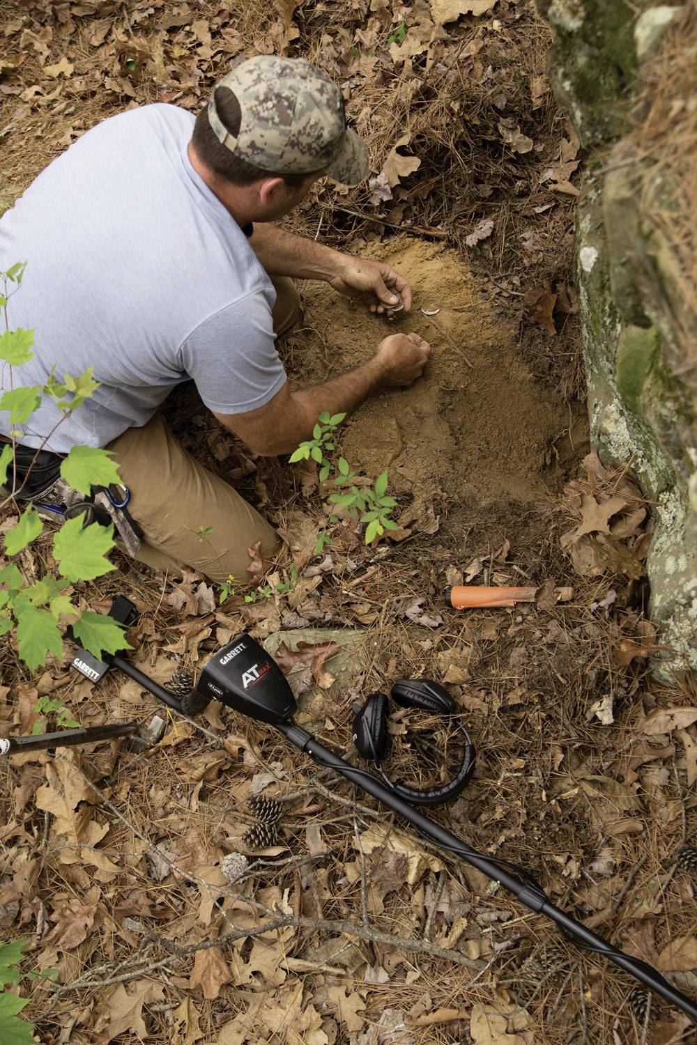 Ein Mann kniet im Wald und gräbt mit den Händen. Neben ihm liegt ein Metalldetektor. Laub und Erde bedecken den Boden.