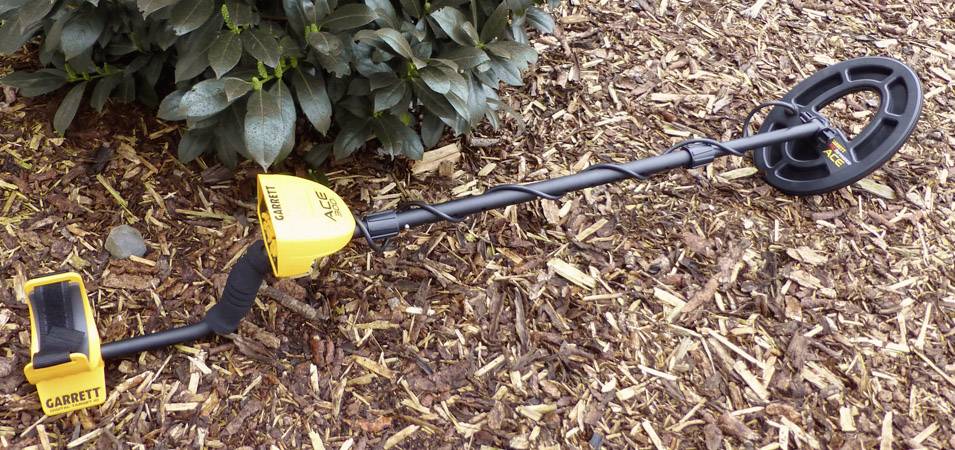 Ein Metalldetektor liegt auf Holzschnitzeln, teilweise vor einem Busch. Gerät ist schwarz-gelb, mit Markenname 'Garrett' sichtbar.