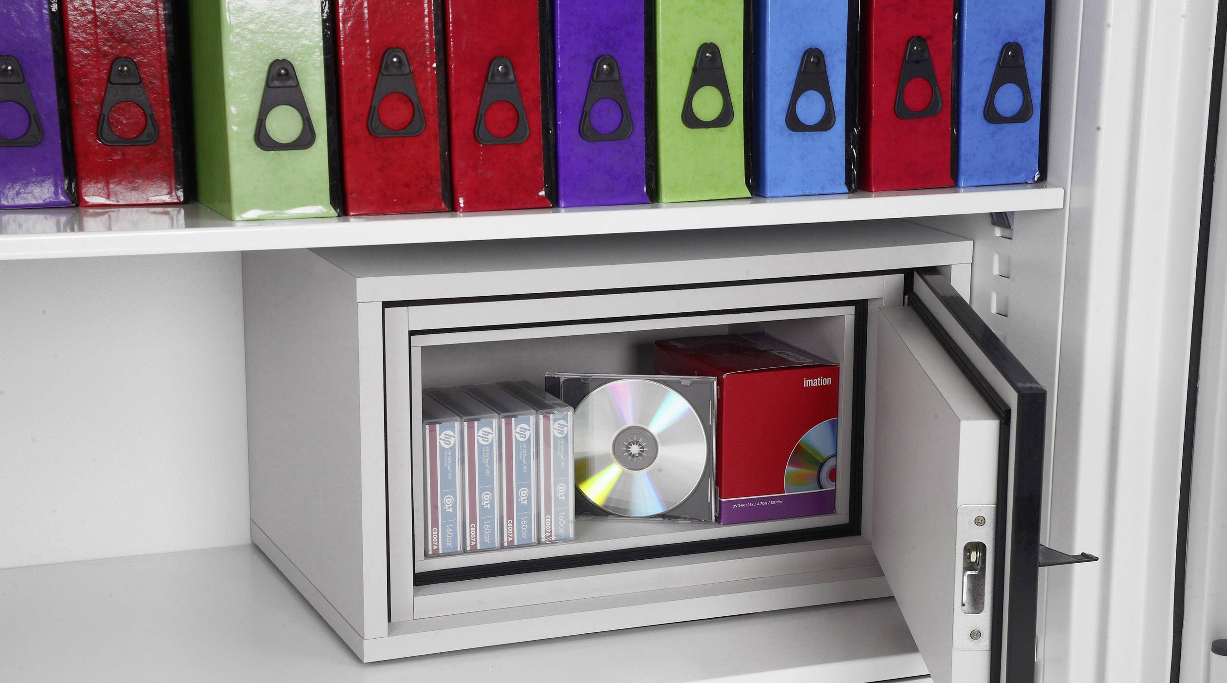 Ein geöffneter Safe in einem Schrank, gefüllt mit CDs und VHS-Kassetten. Darüber befinden sich Ordner in verschiedenen Farben.