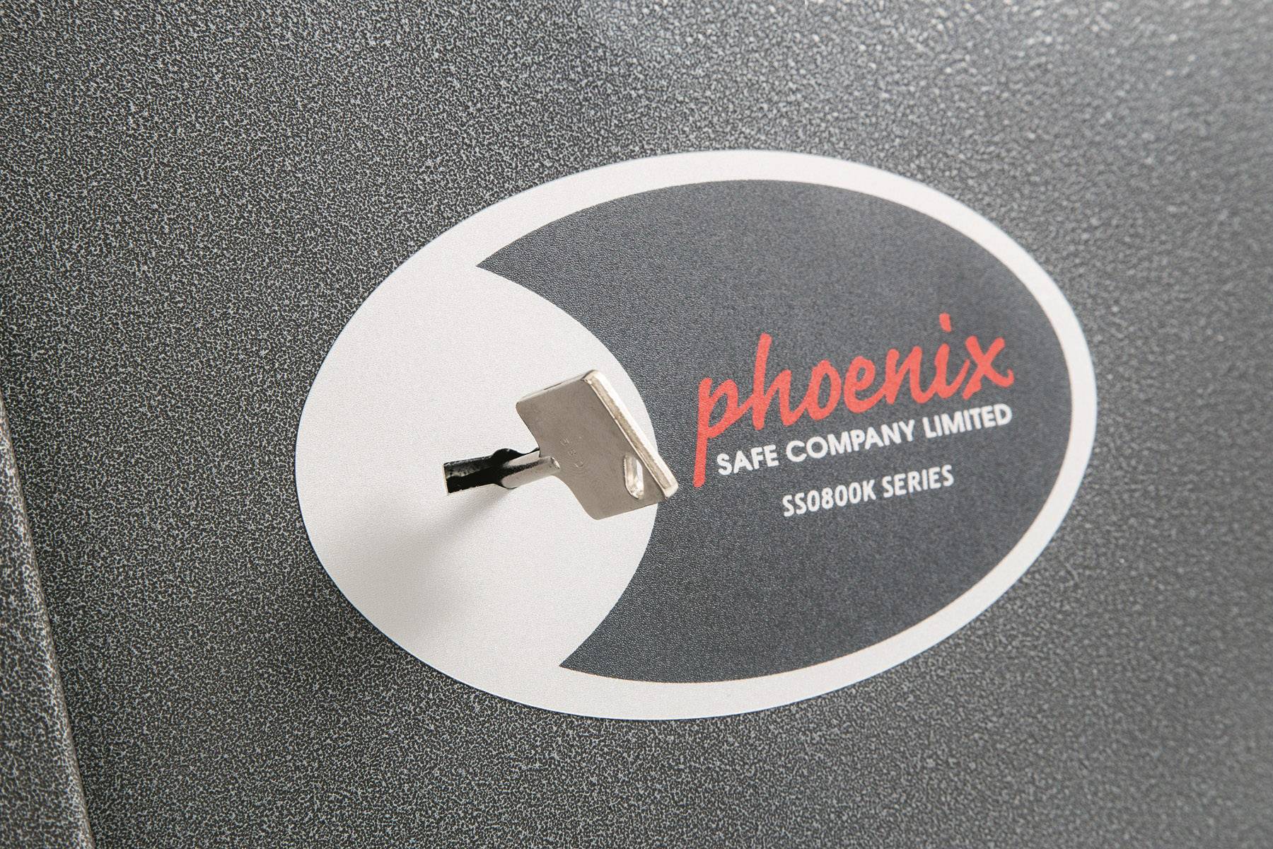Ein Safe von 'Phoenix Safe Company Limited', Modell SS0800K Serie, mit einem Schlüssel im Schloss auf grauer Oberfläche.