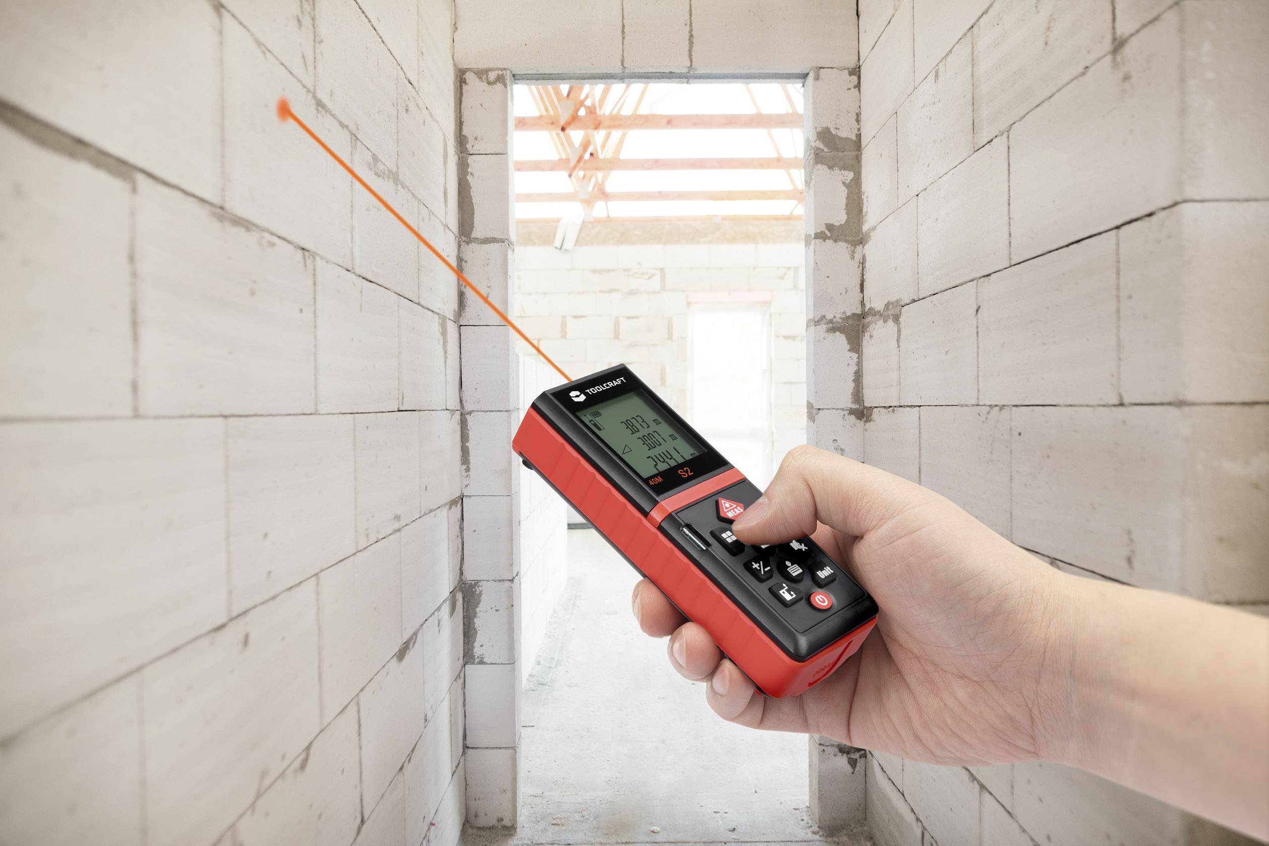Eine Hand hält ein rotes Laser-Entfernungsmessgerät in einem Gebäude im Bau. Ein Laserstrahl richtet sich auf eine Ziegelwand.