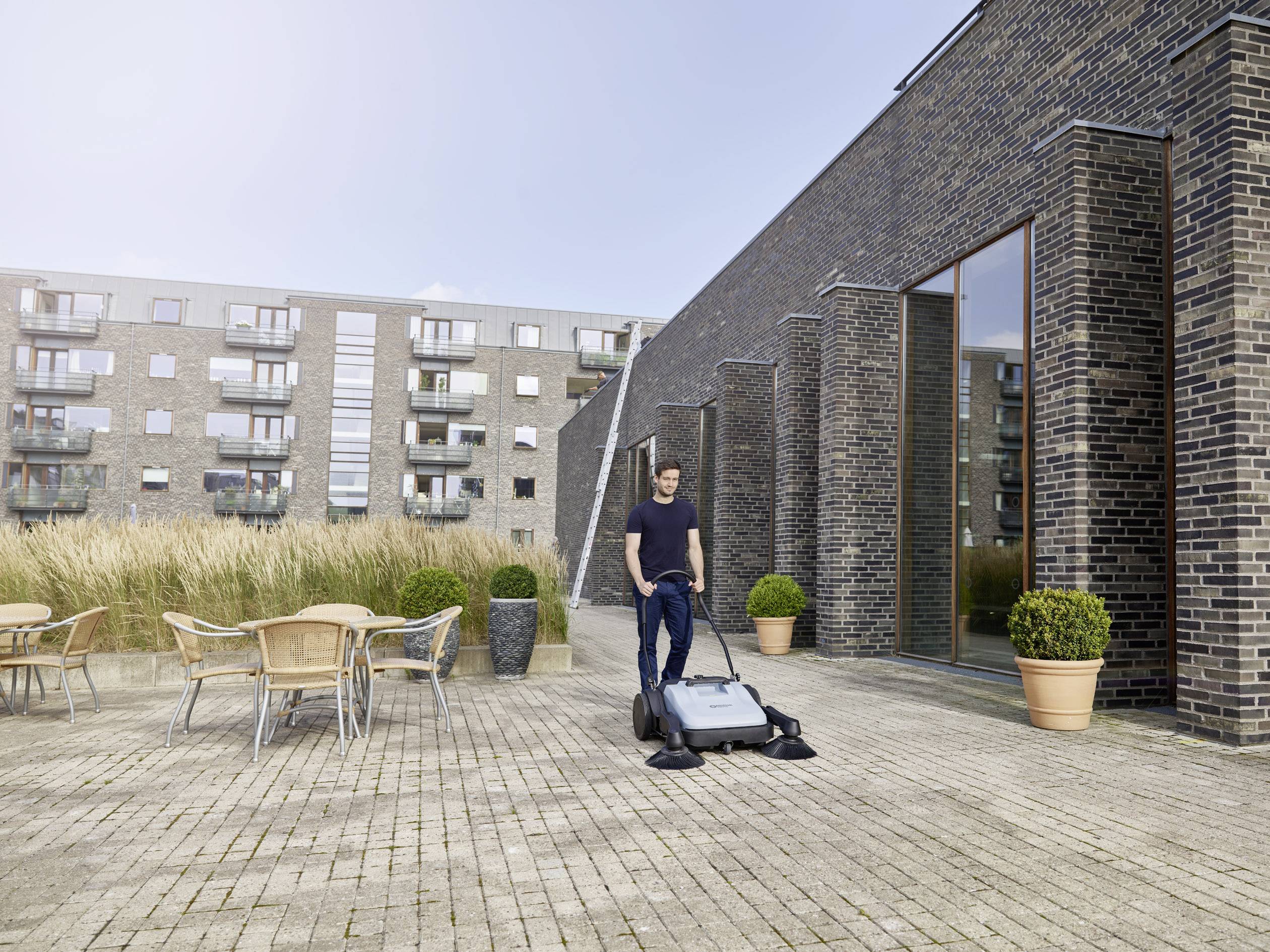Ein Mann benutzt eine große Kehrmaschine auf einer gepflasterten Terrasse neben einem modernen Gebäude mit Glasfenstern und Pflanzen.