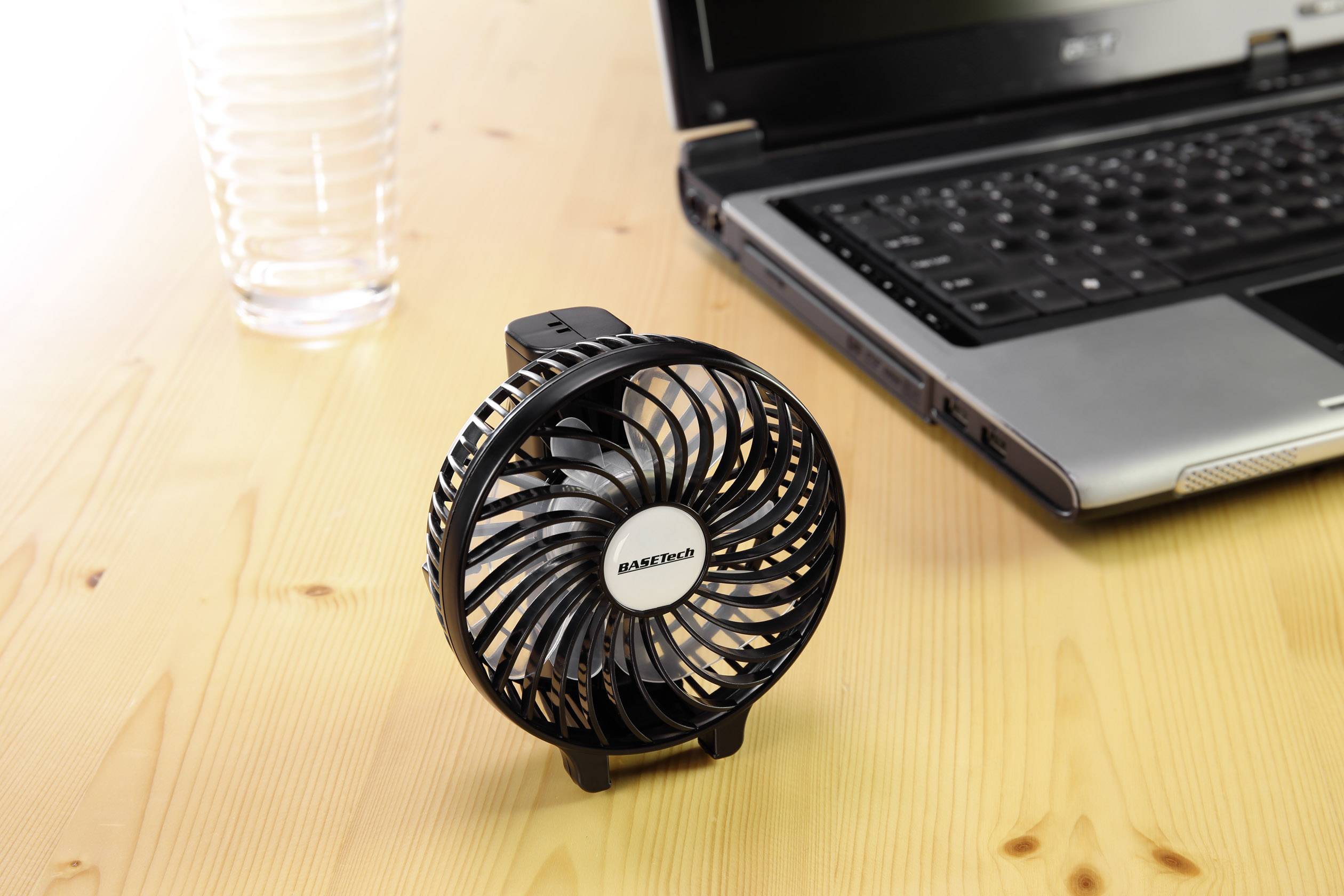 Ein kleiner, schwarzer Tischventilator steht auf einem Holztisch neben einem halb gefüllten Glas und einem aufgeklappten Laptop.