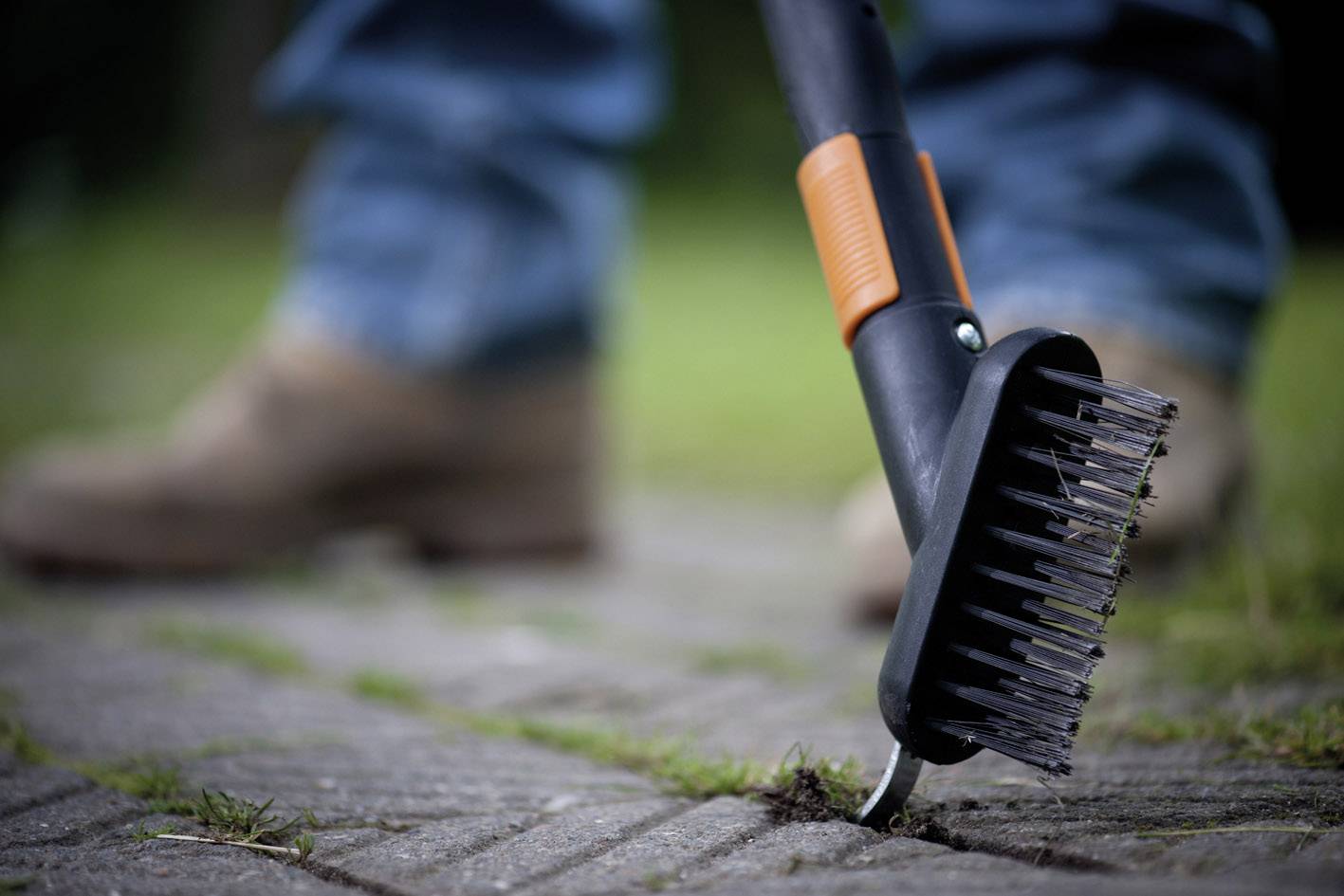Eine Person mit blauen Jeans entfernt Unkraut mit einem Unkrautkratzer aus Fugen auf einer gepflasterten Fläche. Im Hintergrund sind verschwommen grüne Pflanzen erkennbar.