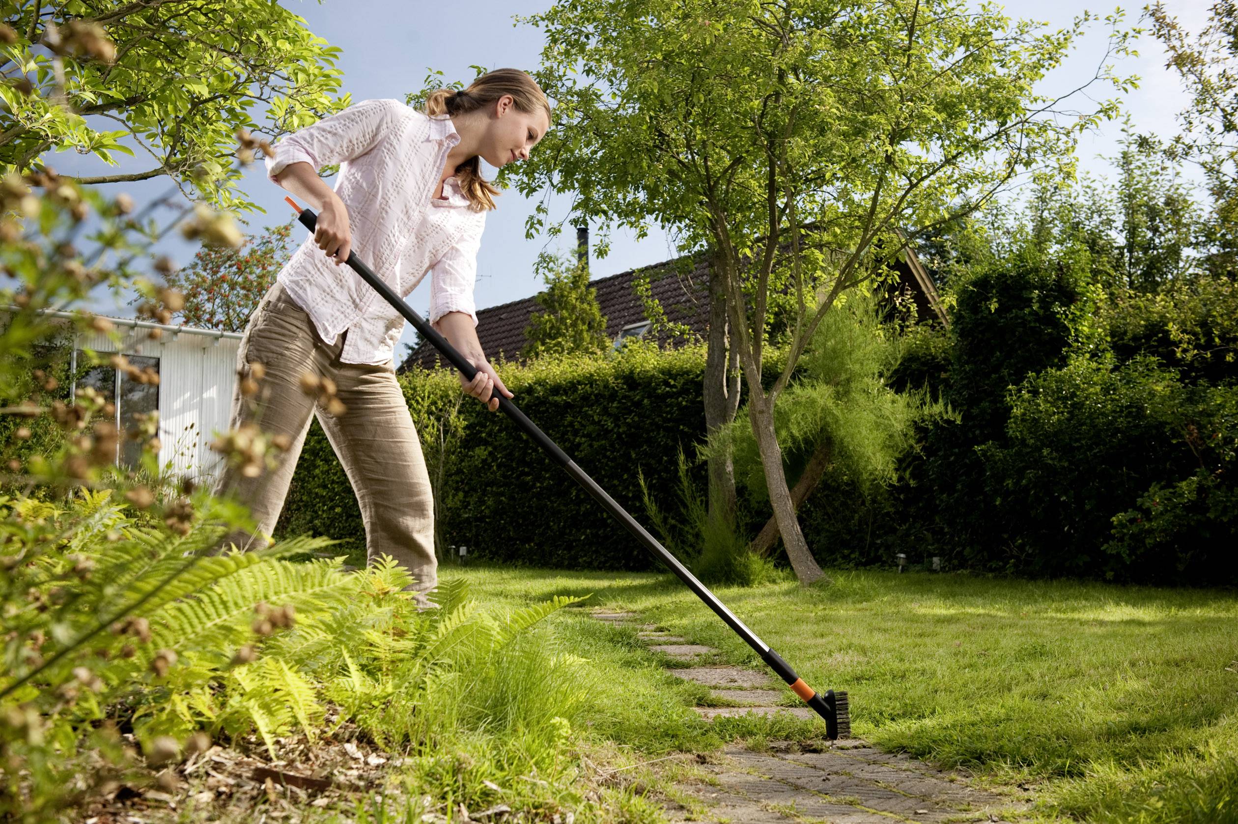 Person trimmt Rasen neben einem Gartenpfad mit einem Kantenschneider. Im Hintergrund sind Bäume und Sträucher.