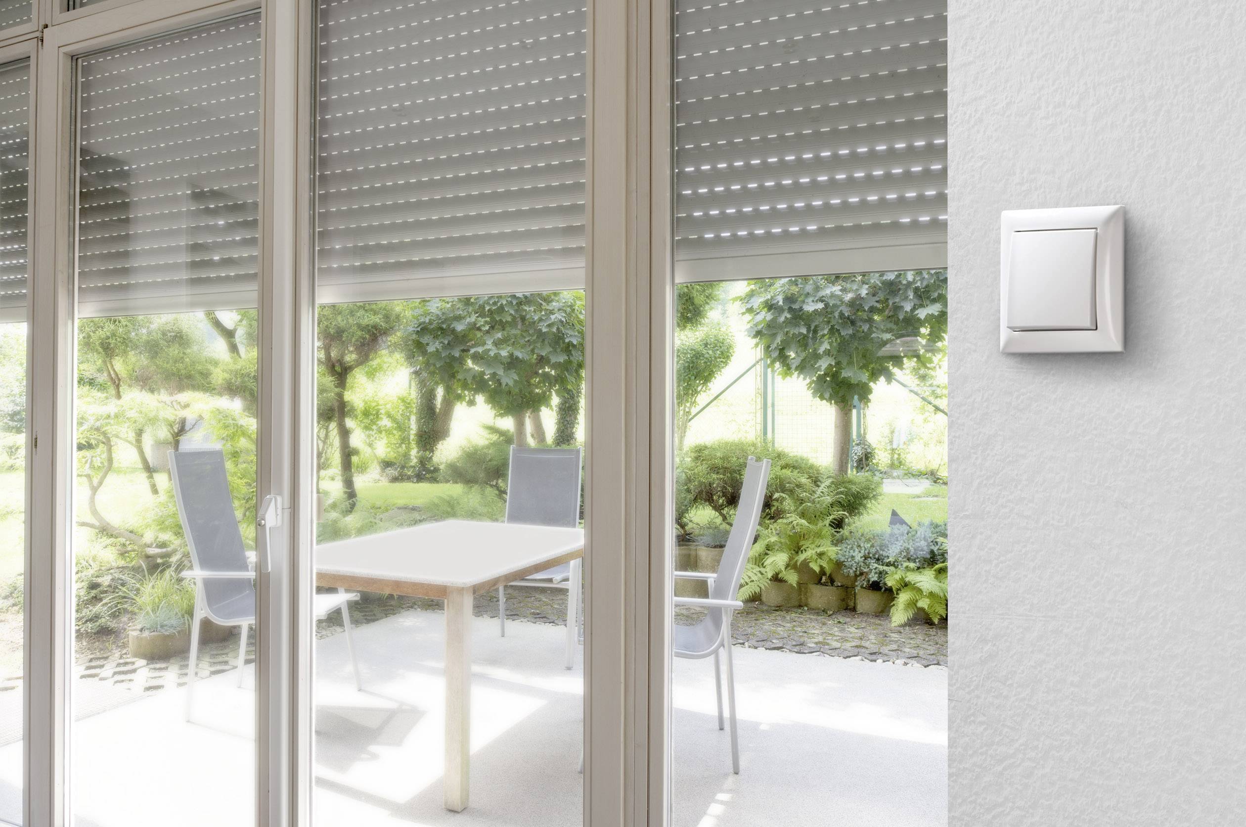 Eine Terrasse mit Tisch und Stühlen, sichtbar durch eine Glastür mit heruntergelassenen Rollläden. Ein Schalter an der Wand rechts.