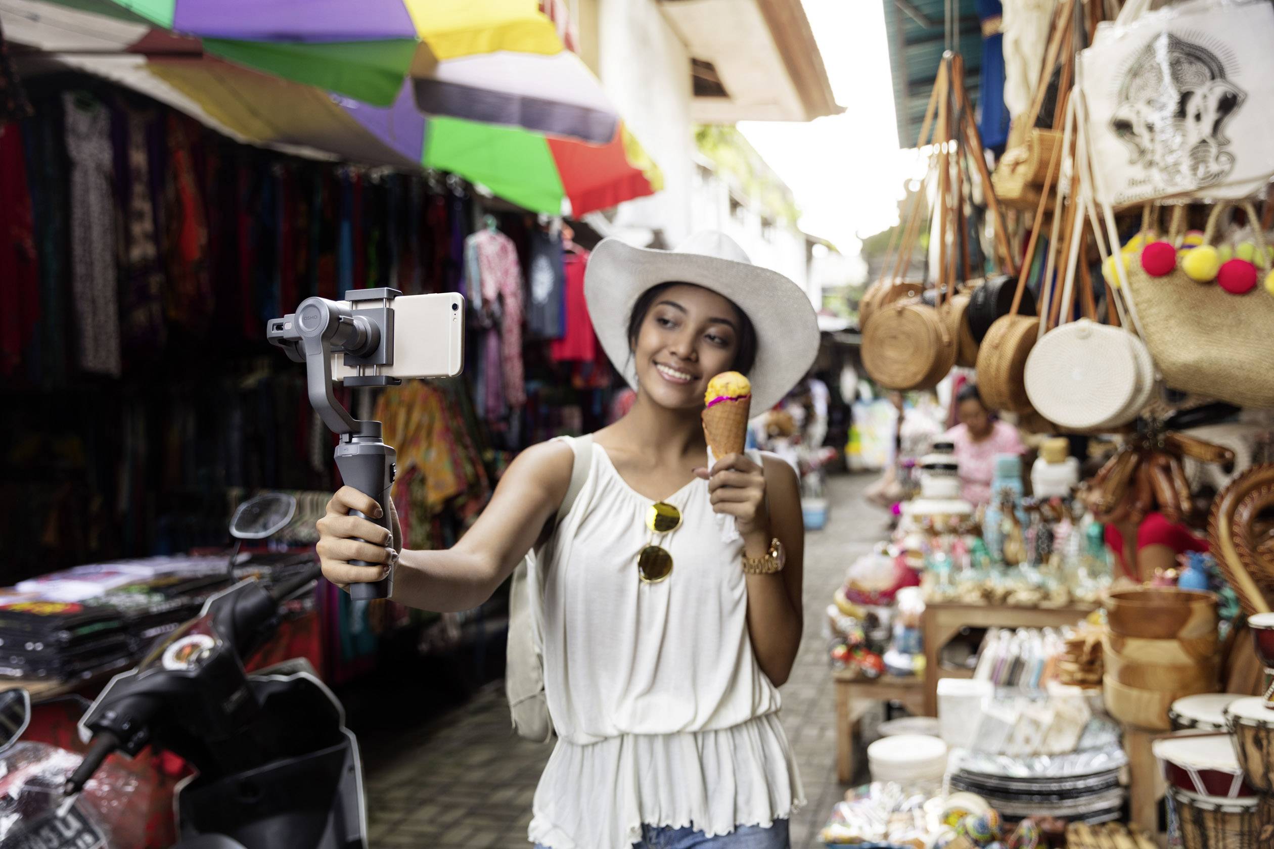 Person in Hut isst Eis und macht Selfie im belebten Markt mit bunten Ständen voller Waren im Hintergrund.