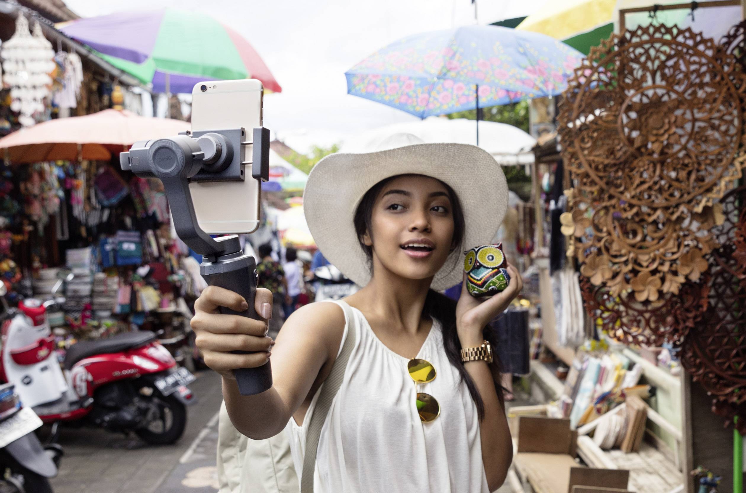 Frau in Sommerkleidung und Hut macht ein Selfie mit einem Smartphone in einer belebten Marktumgebung, hält bunte Figur in der Hand.