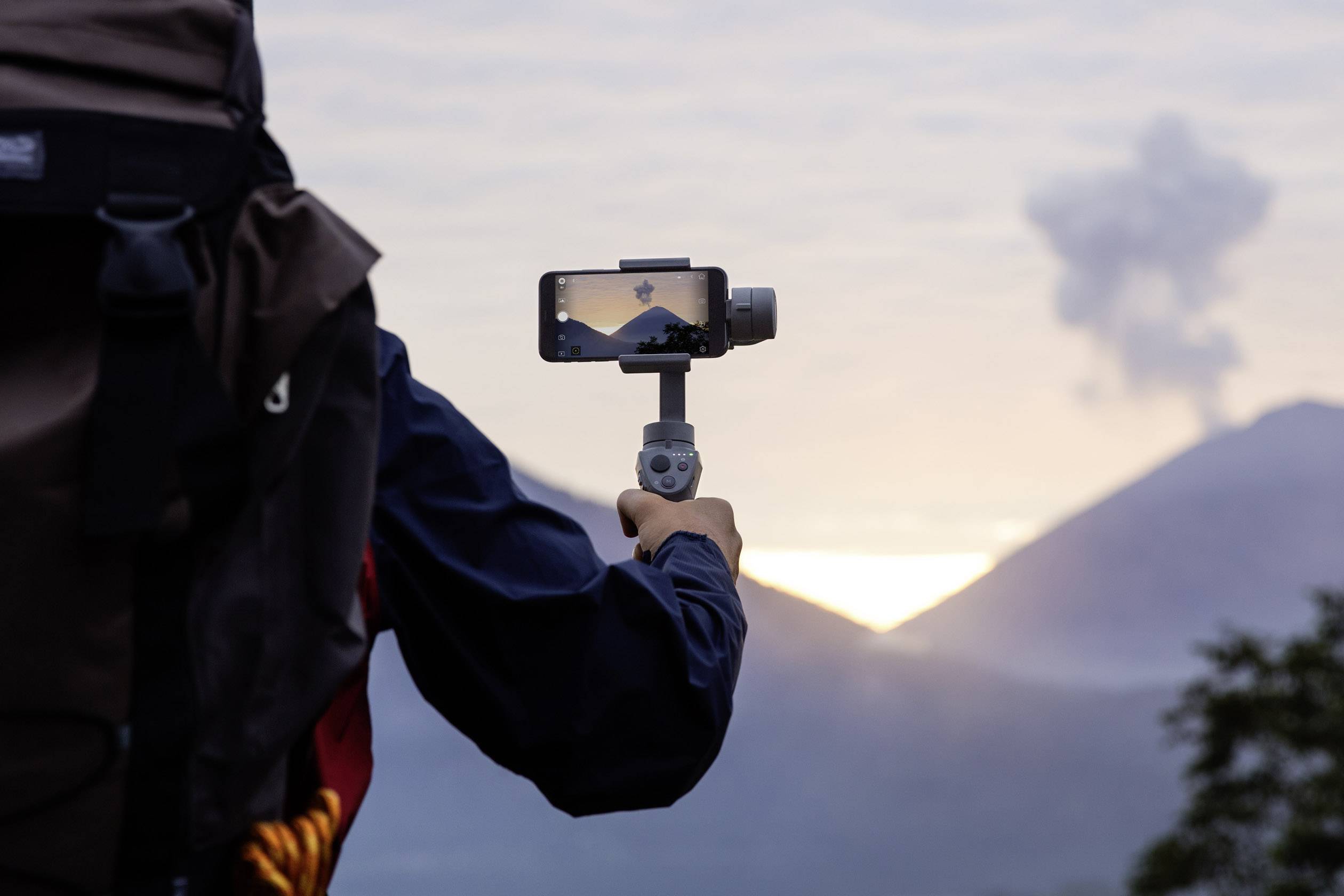 Eine Person fotografiert mit einem Smartphone an einem Gimbal eine Berglandschaft im Sonnenaufgang, mit Vulkanrauch im Hintergrund.