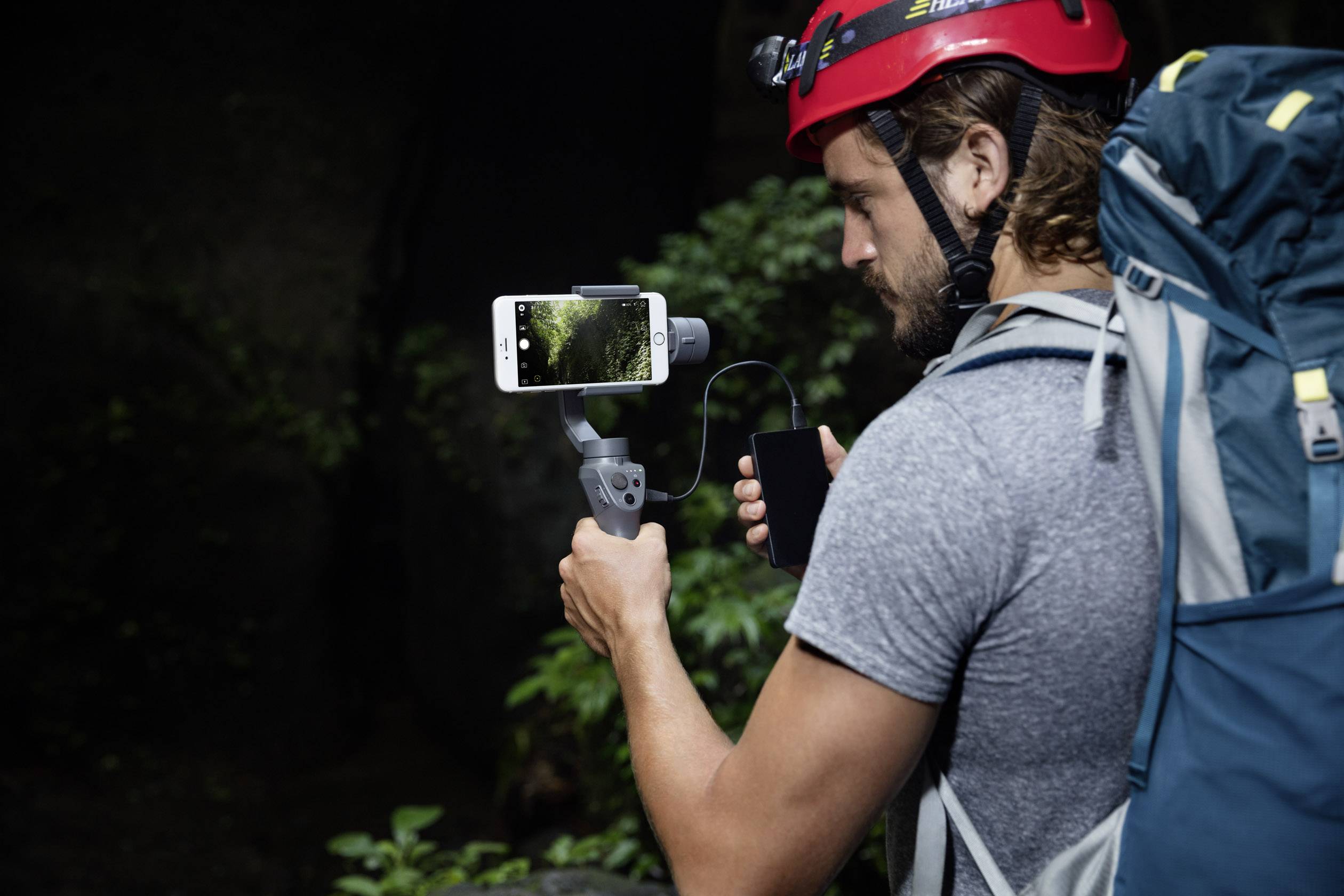 Eine Person mit rotem Helm und Rucksack filmt mit einem Smartphone auf einem Stabilisator in einem bewaldeten Umfeld.