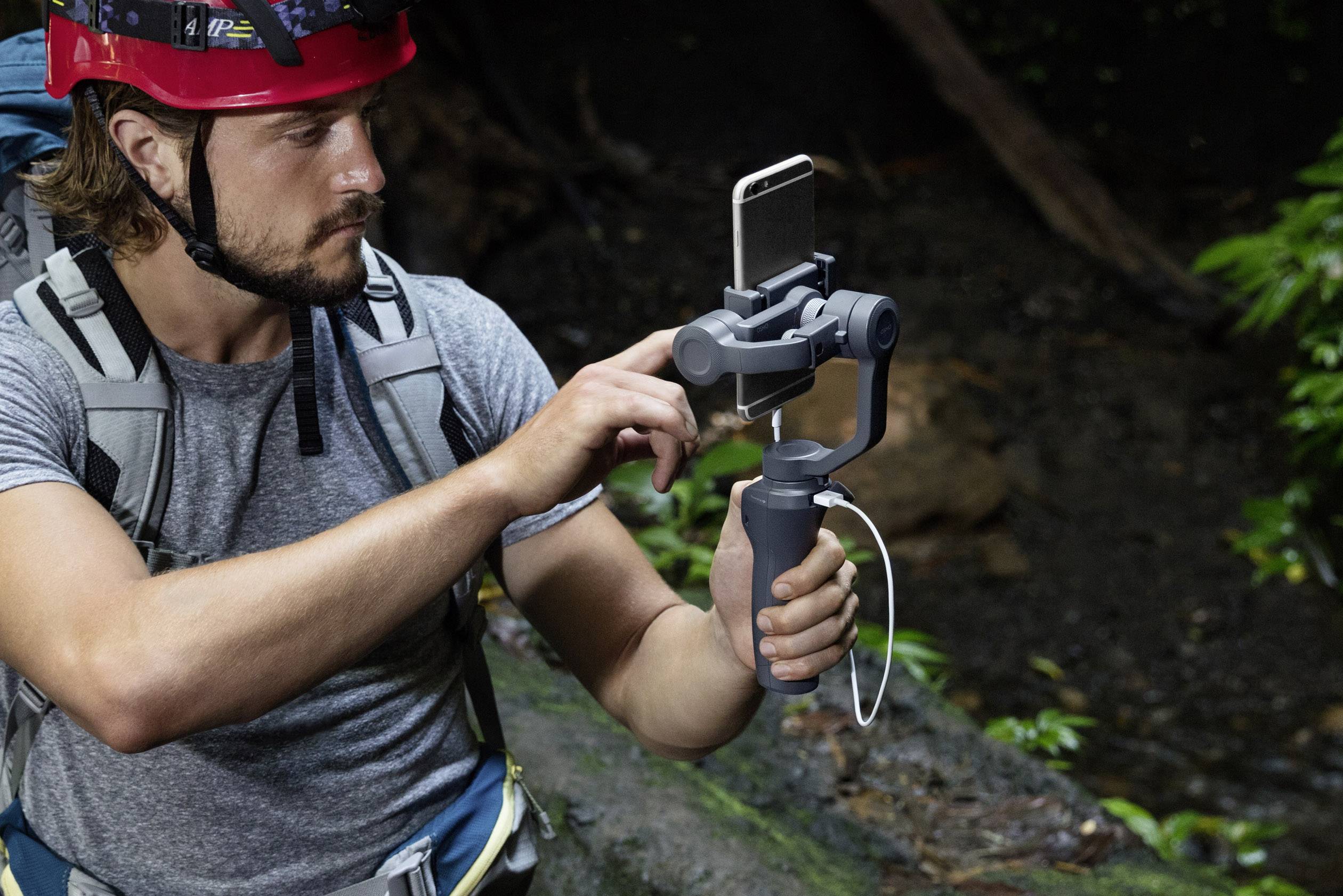 Ein Mann im Wald, trägt einen roten Helm und Rucksack. Er bedient ein Smartphone auf einem Stabilisator, vermutlich für Videoaufnahmen.