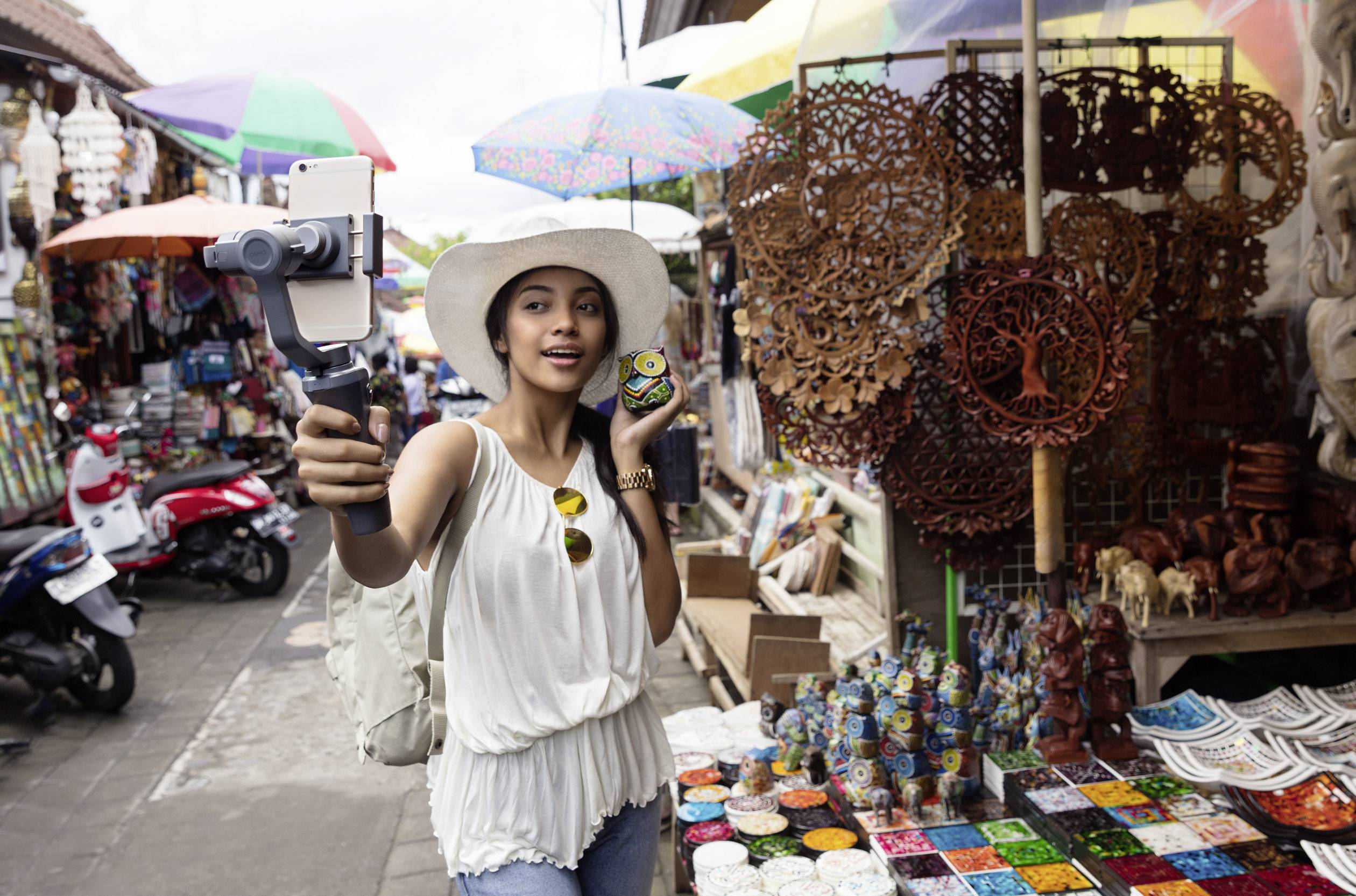 Eine Frau mit Hut macht ein Selfie auf einem Marktstand. Sie hält eine bunte Kugel in der Hand. Im Hintergrund sind Souvenirs und bunte Regenschirme.