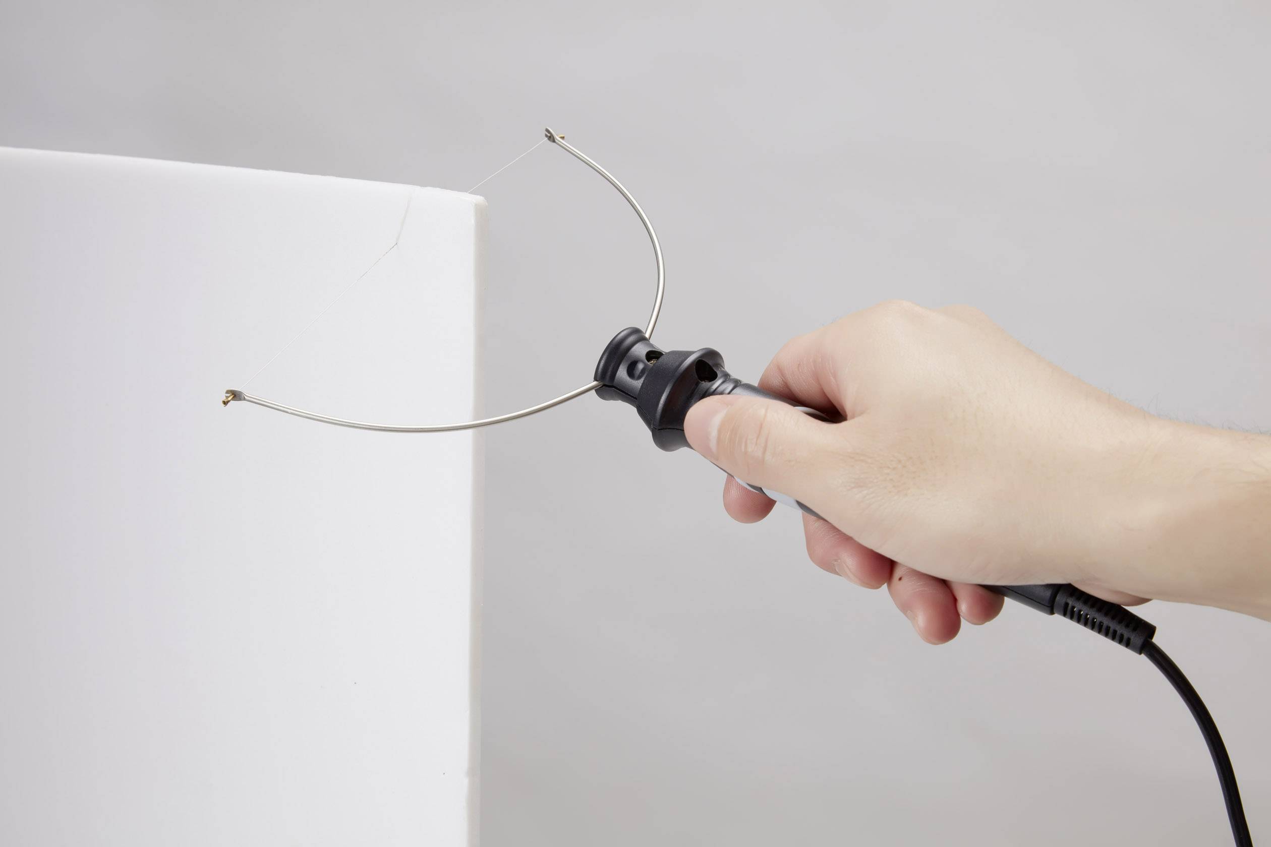 Eine Hand hält ein Werkzeug mit gebogenem Draht, das auf einen weißen Block gerichtet ist, vermutlich zum Schneiden oder Formen.