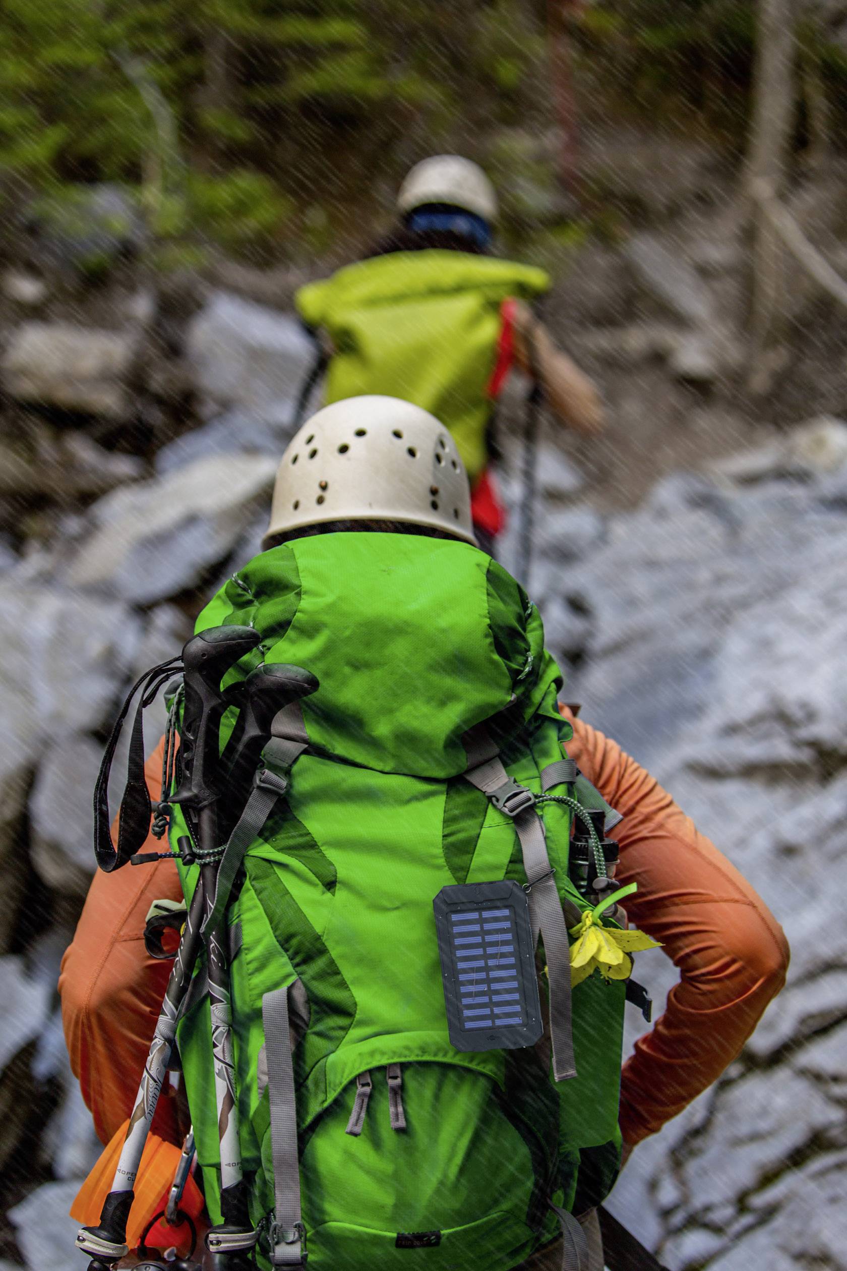Zwei Personen klettern auf einem felsigen Pfad. Die vordere Person trägt einen grünen Rucksack und einen Helm. Bäume im Hintergrund.