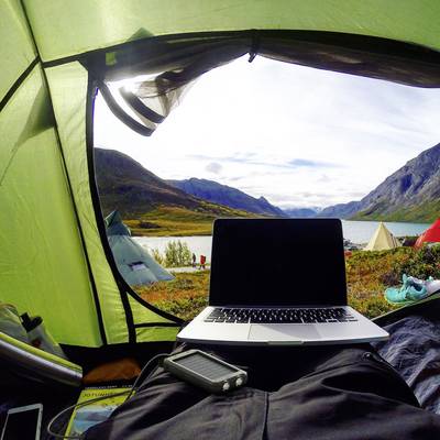 Eine Person liegt im Zelt mit einem Laptop auf dem Schoß, mit Blick auf eine malerische Berg- und Seelandschaft im Hintergrund.