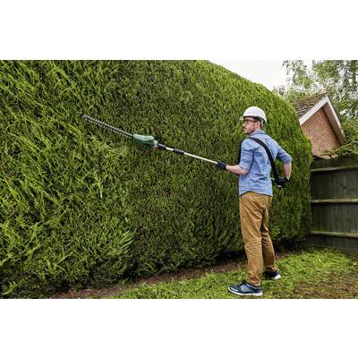 Ein Mann mit Schutzhelm und Handschuhen schneidet eine große Hecke mit einem elektrischen Heckenschneider in einem Garten.