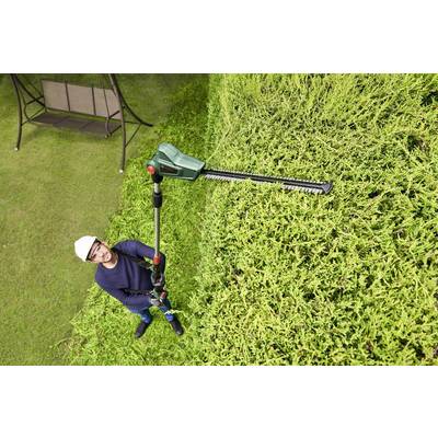 Eine Person in Schutzkleidung schneidet mit einer elektrischen Heckenschere die Oberseite einer hohen Hecke.