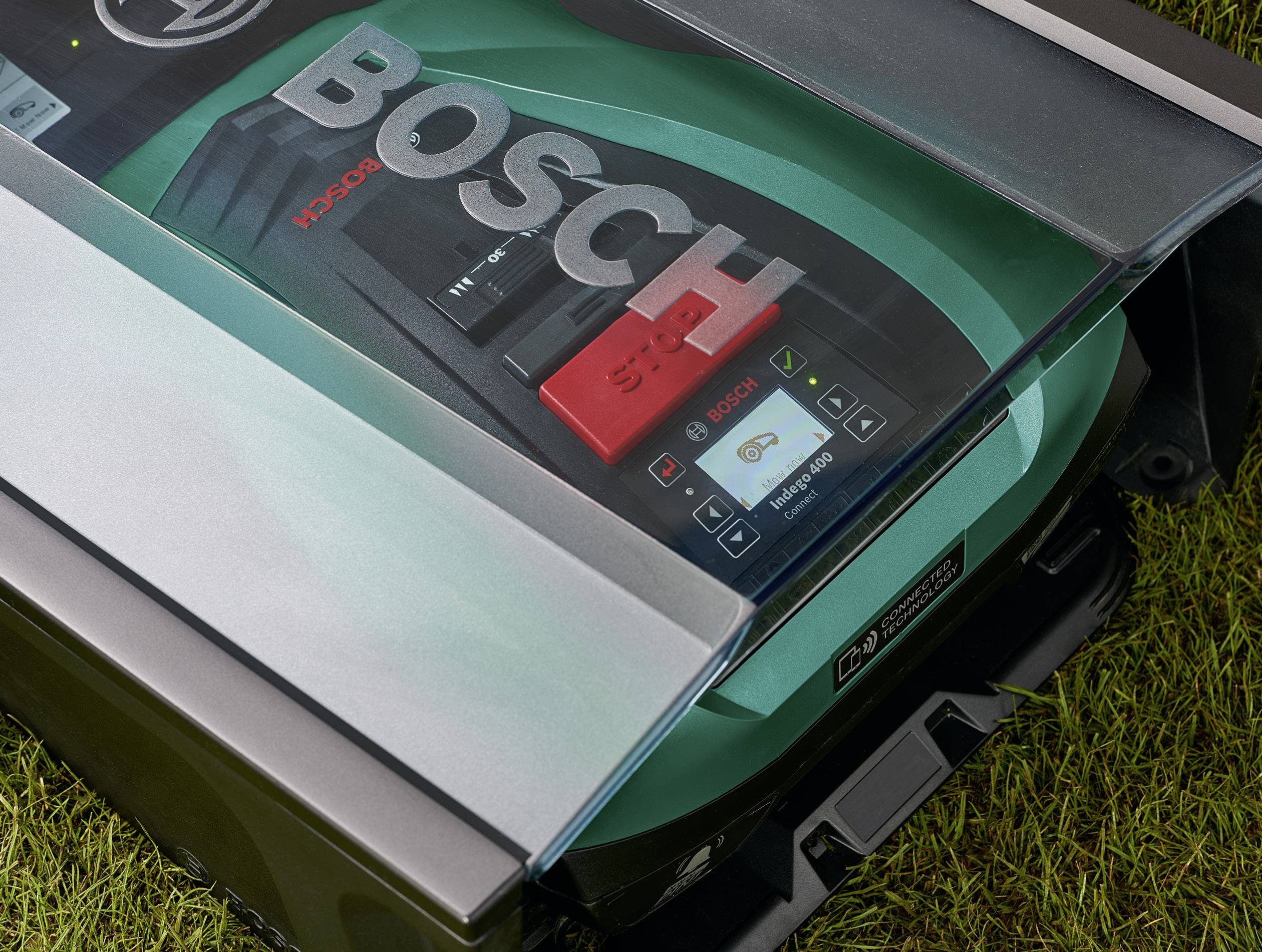 Rasenmäher mit 'BOSCH'-Aufschrift auf grünem Gehäuse, unter transparentem Schutzdeckel, auf Rasenfläche. Digitale Anzeige sichtbar.