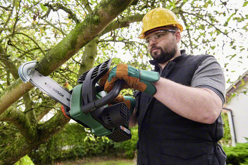 Ein Mann in Arbeitskleidung und Schutzhelm schneidet mit einer elektrischen Kettensäge einen Ast von einem Baum ab.