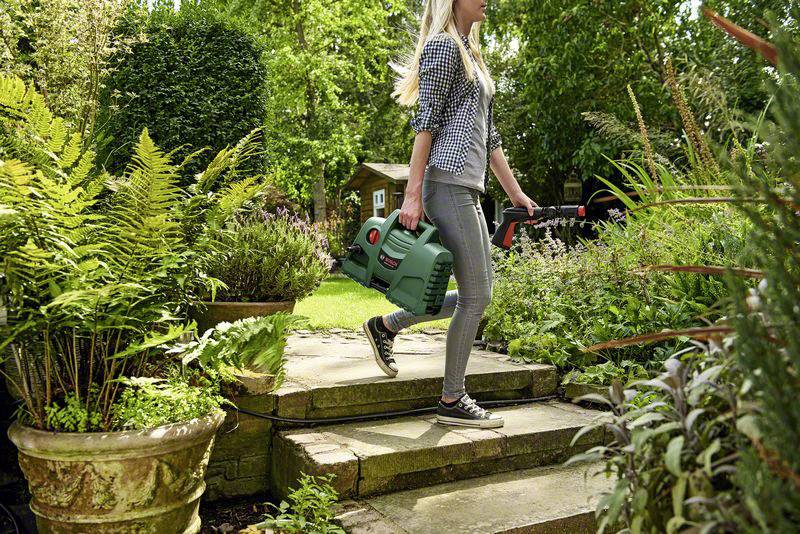 Eine Person trägt ein tragbares Reinigungsgerät durch einen grünen Garten mit verschiedenen Pflanzen und Töpfen.