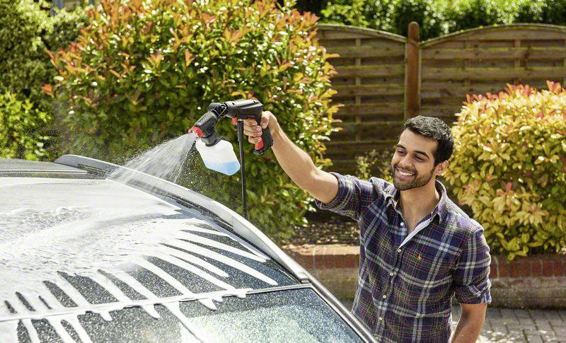 Mann wäscht ein Auto mit einer Schaumsprühpistole in einem Gartenbereich an einem sonnigen Tag, während er lächelt.