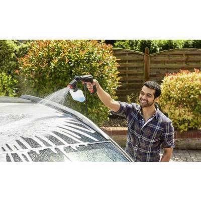 Mann wäscht ein Auto mit einer Schaumsprühpistole in einem Gartenbereich an einem sonnigen Tag, während er lächelt.