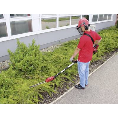 Eine Person in Schutzkleidung schneidet mit einer elektrischen Heckenschere Büsche entlang eines Gebäudes.