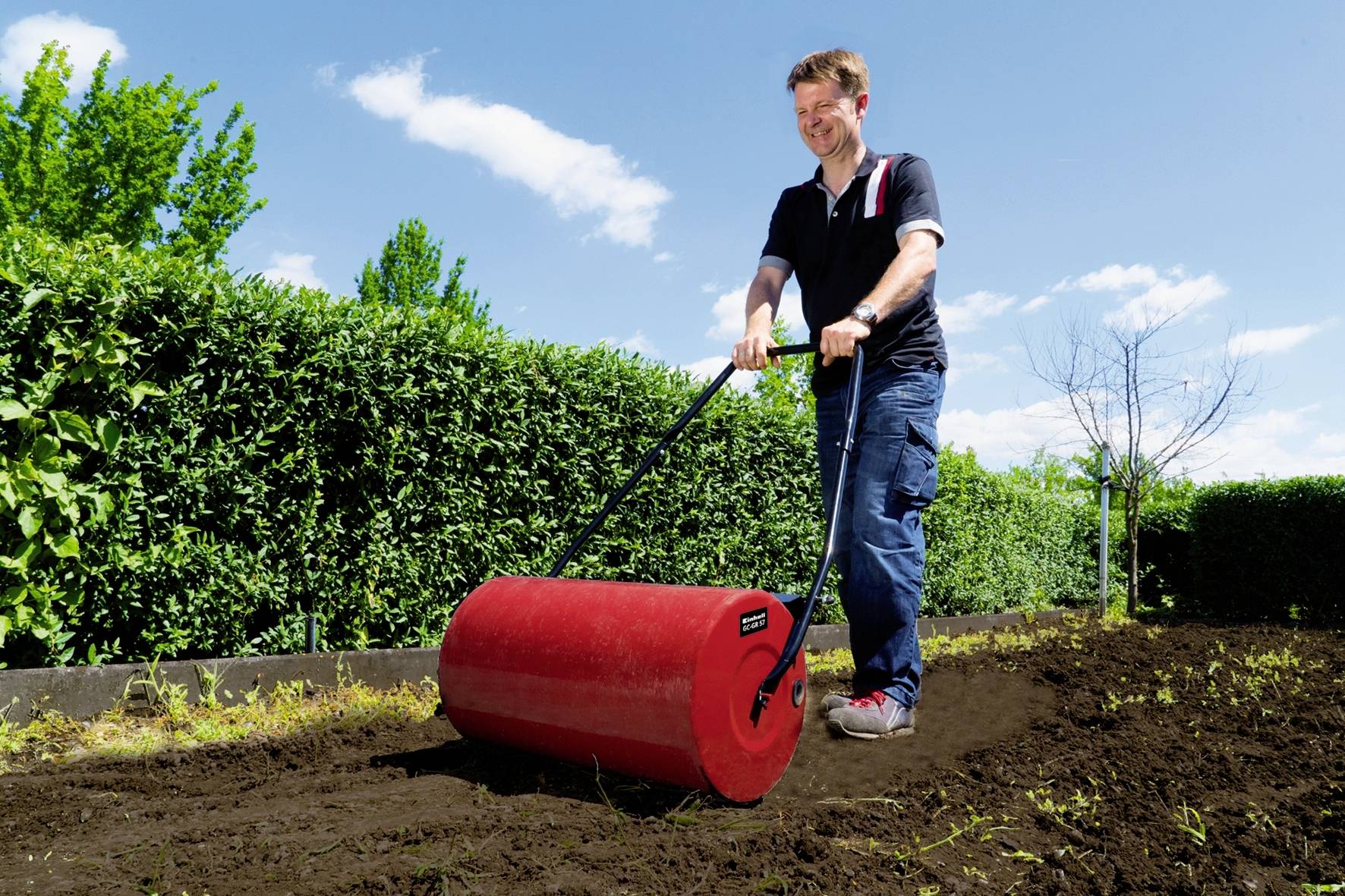 Eine Person schiebt einen roten Gartenroller über frisch aufgelockerte Erde in einem Garten, umgeben von grünen Hecken unter blauem Himmel.