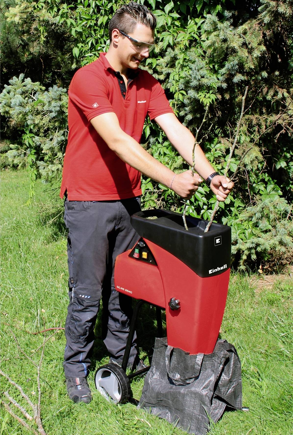 Ein Mann in rotem Shirt benutzt einen roten Gartenhäcksler im Freien, um Äste zu zerkleinern. Er steckt Zweige in das Gerät.