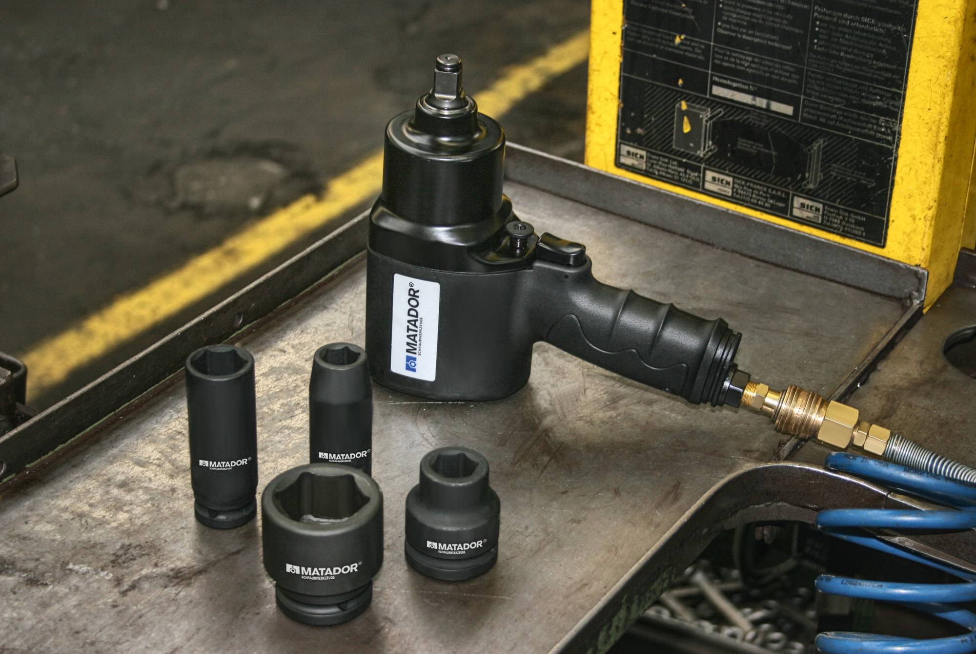 Luftschlagschrauber und fünf Stecknüsse auf einer Metallwerkbank in einer Werkstatt. Im Hintergrund Werkstattzubehör und gelbe Linie.