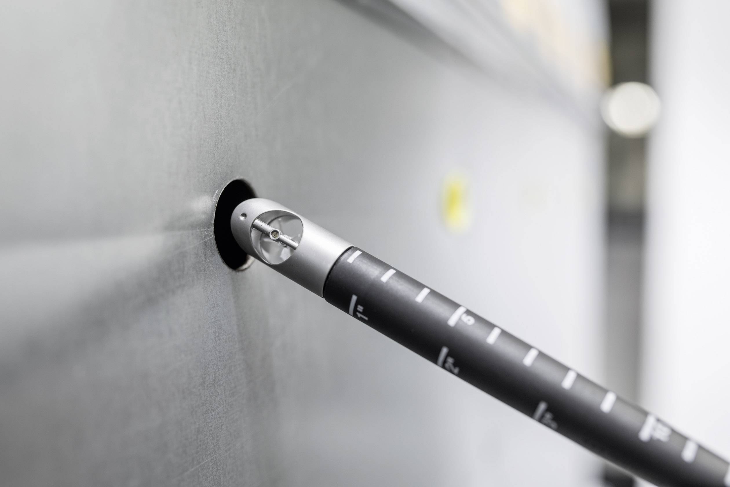 Ein metallischer Stab mit Skalenmarkierungen ist schräg in eine graue Wand eingelassen, möglicherweise ein Messinstrument oder Werkzeug.