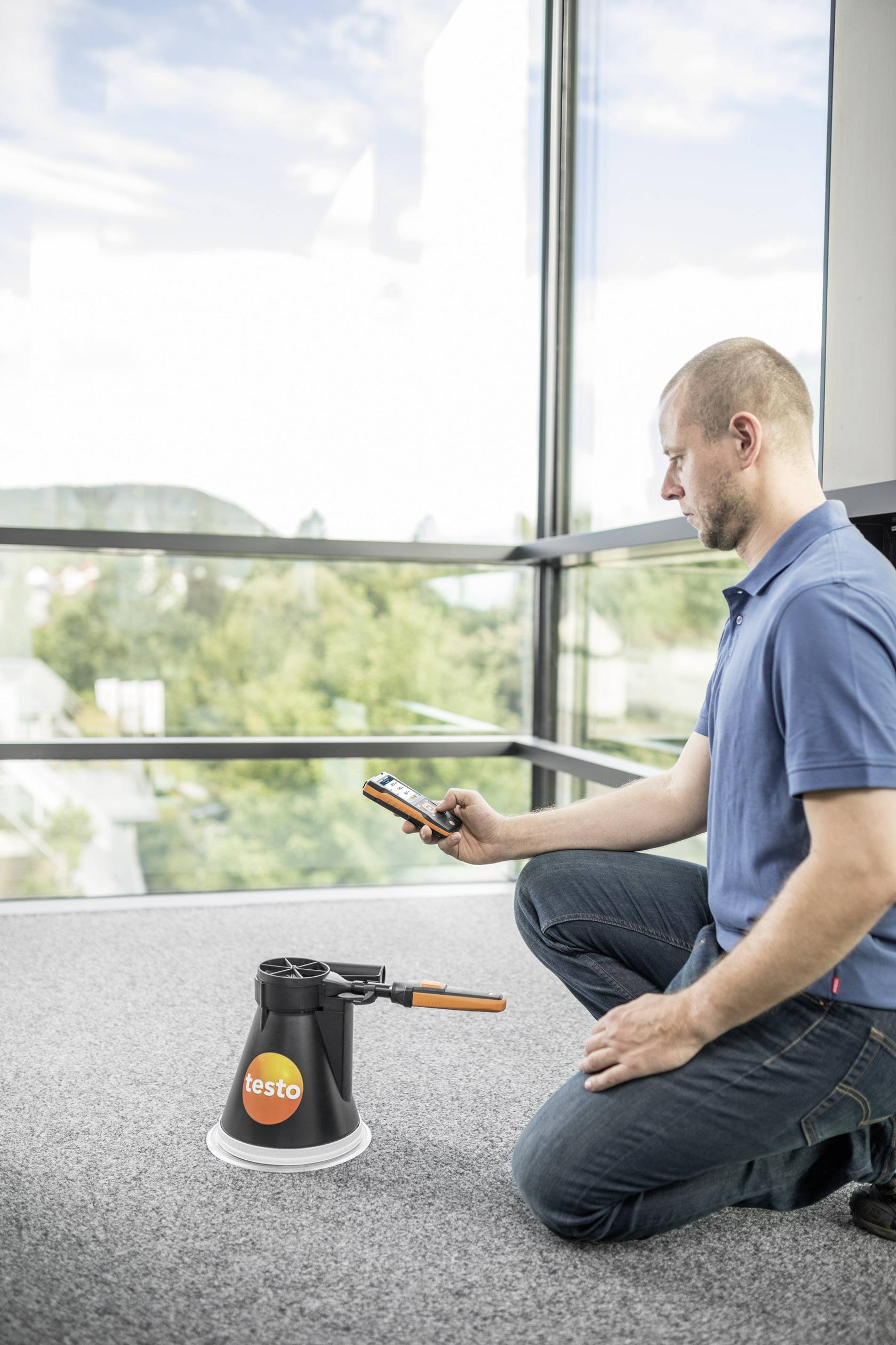 Ein Mann kniet auf einem Teppichboden neben einem schwarzen Messgerät und hält ein Handy, während er auf einen Bildschirm schaut.