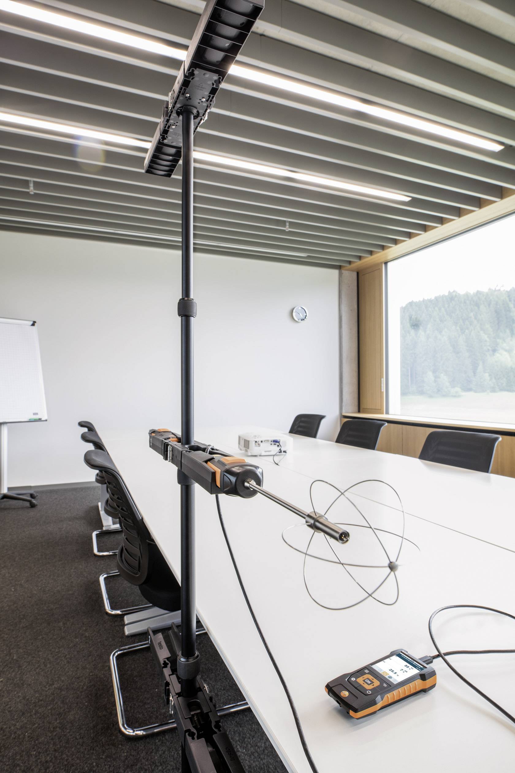 Konferenzraum mit langem Tisch, sieben Stühlen, Flipchart. Im Vordergrund ein Messgerät auf einem Stativ mit Antenne. Große Fenster mit Blick ins Grüne.