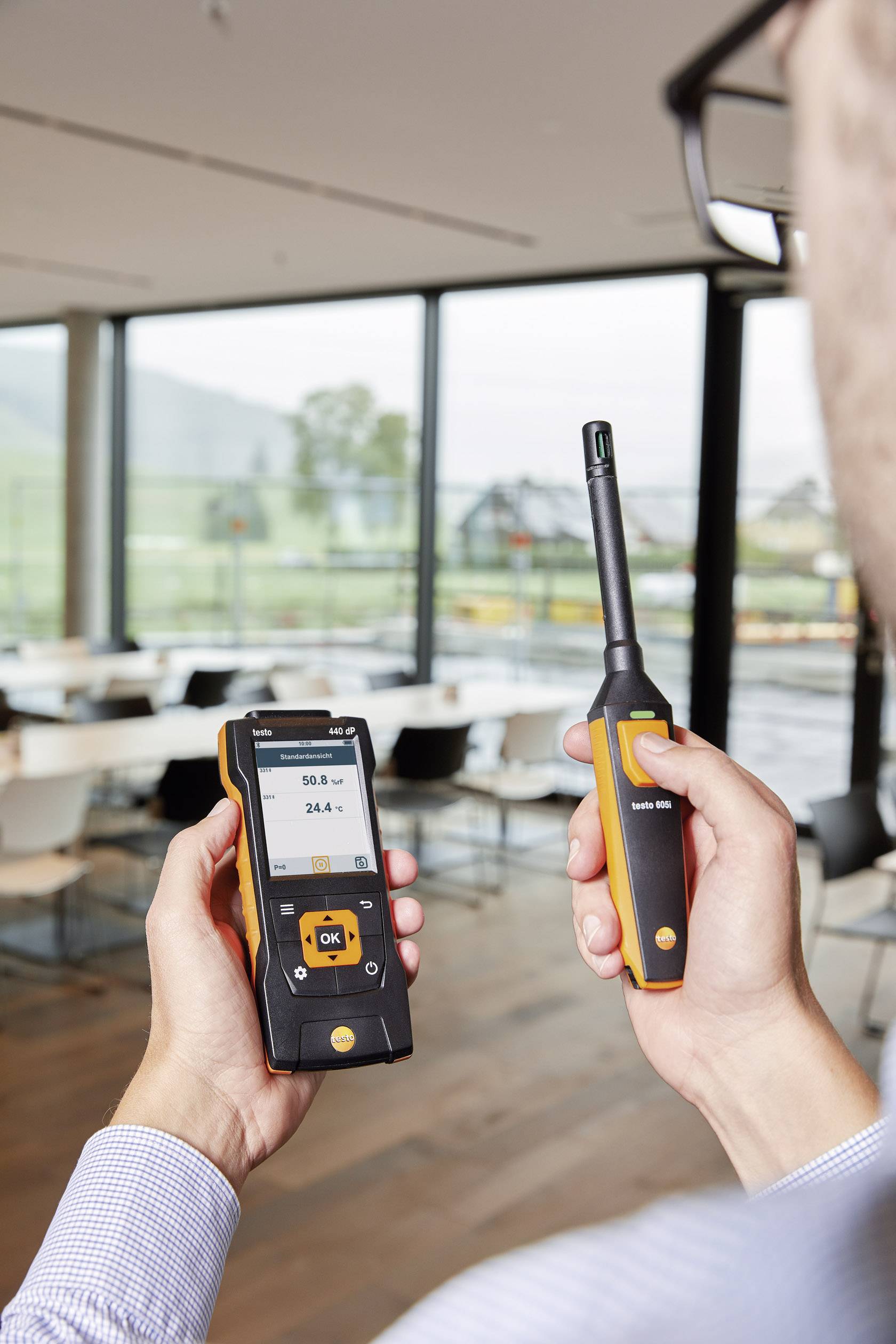 Eine Person hält ein digitales Messgerät in der Hand, das Luftqualität misst. Es zeigt Daten auf einem Display. Im Hintergrund sind Fenster und ein Tisch.